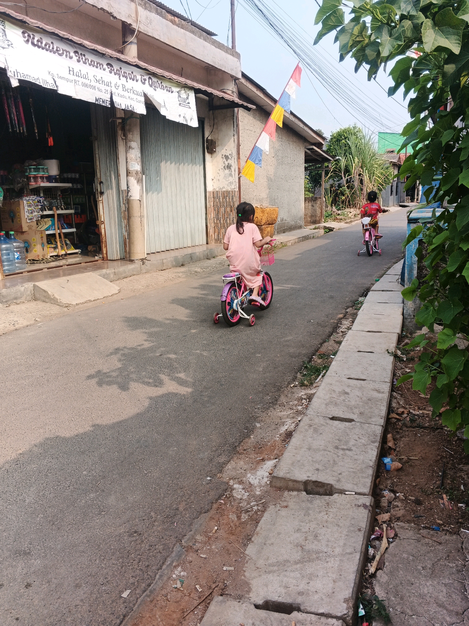 In the city of Kadu, Indonesia, two young girls ride bicycles down a paved street lined with simple buildings and dense foliage. The girl in the foreground, wearing a pink dress, is on a pink bicycle with training wheels. The second girl, further down the street, wears a red shirt and rides a pink bicycle. A sign above a storefront reads "Riddem Khain Aqiqah & Qurban," with smaller text indicating "Halal, Sehat & Berkualitas" and "Kp. Sempur RT. 006/006 No. 25 Kel. Kadu, Kec. Curug." A collection of flags, red, white, and blue, are strung across wires overhead. The street is partially shaded by trees, and a concrete drainage ditch runs along the right side of the road, filled with some debris. The overall scene depicts a casual, everyday moment in a residential area.