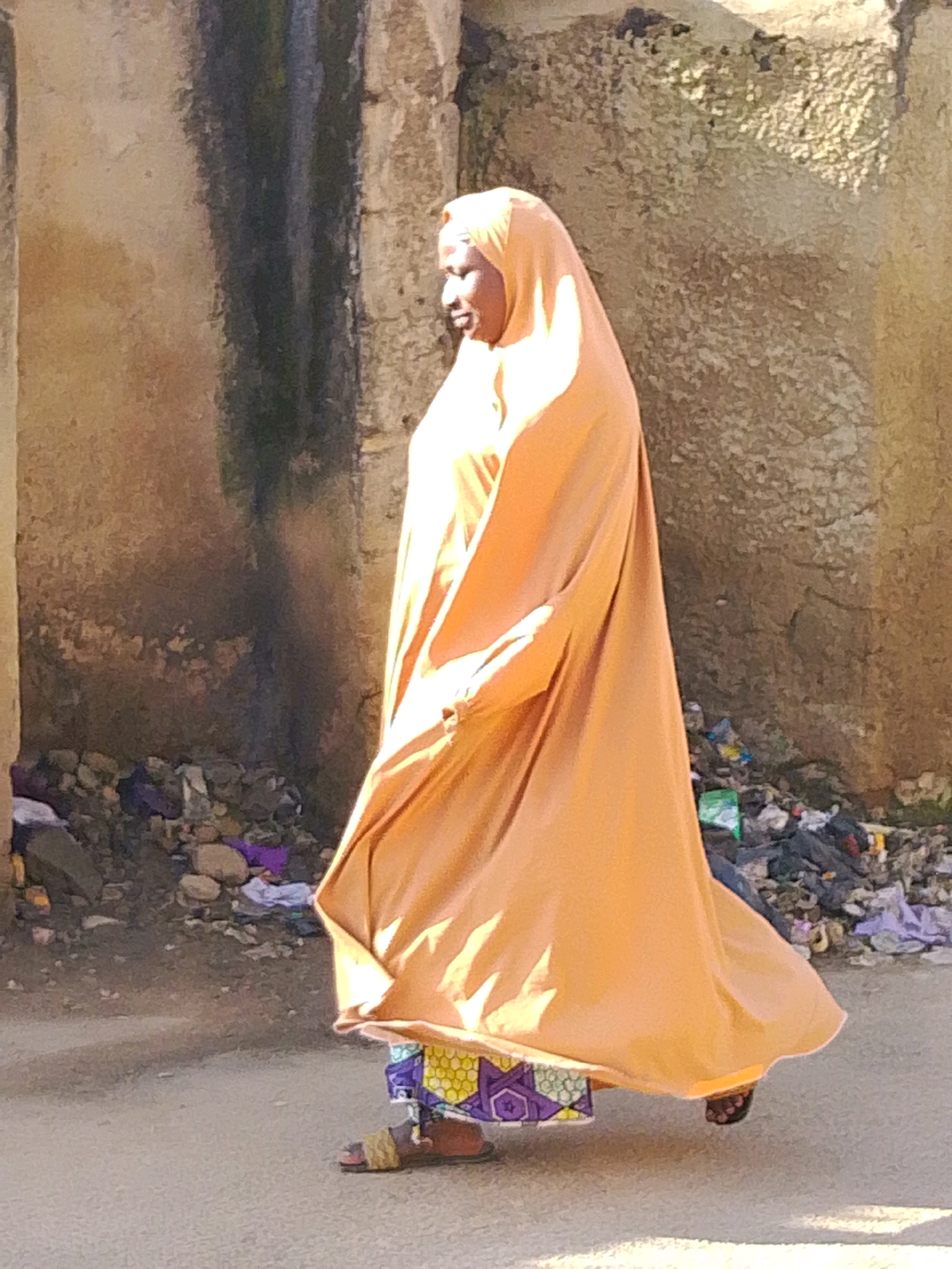 A woman wearing an orange hijab and dress walks on a paved street in Jos, Nigeria. She is also wearing a patterned skirt and sandals. The street is lined with a stone wall and piles of refuse.