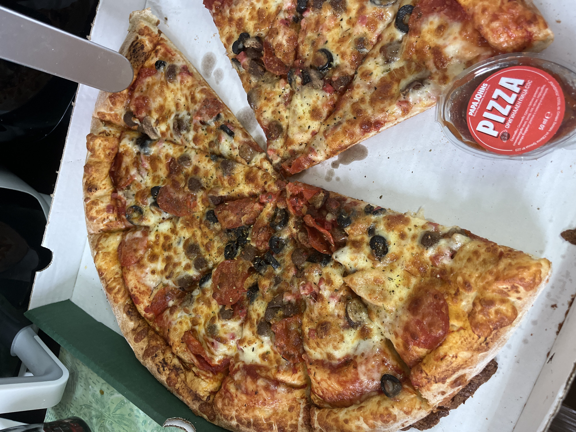 A partially eaten pizza from Papa Johns is in a white pizza box. The pizza is topped with cheese, pepperoni, sausage, mushrooms, and black olives. Next to the pizza is a small container of Papa Johns pizza sauce. A metal spatula is near the pizza. The scene is indoors.