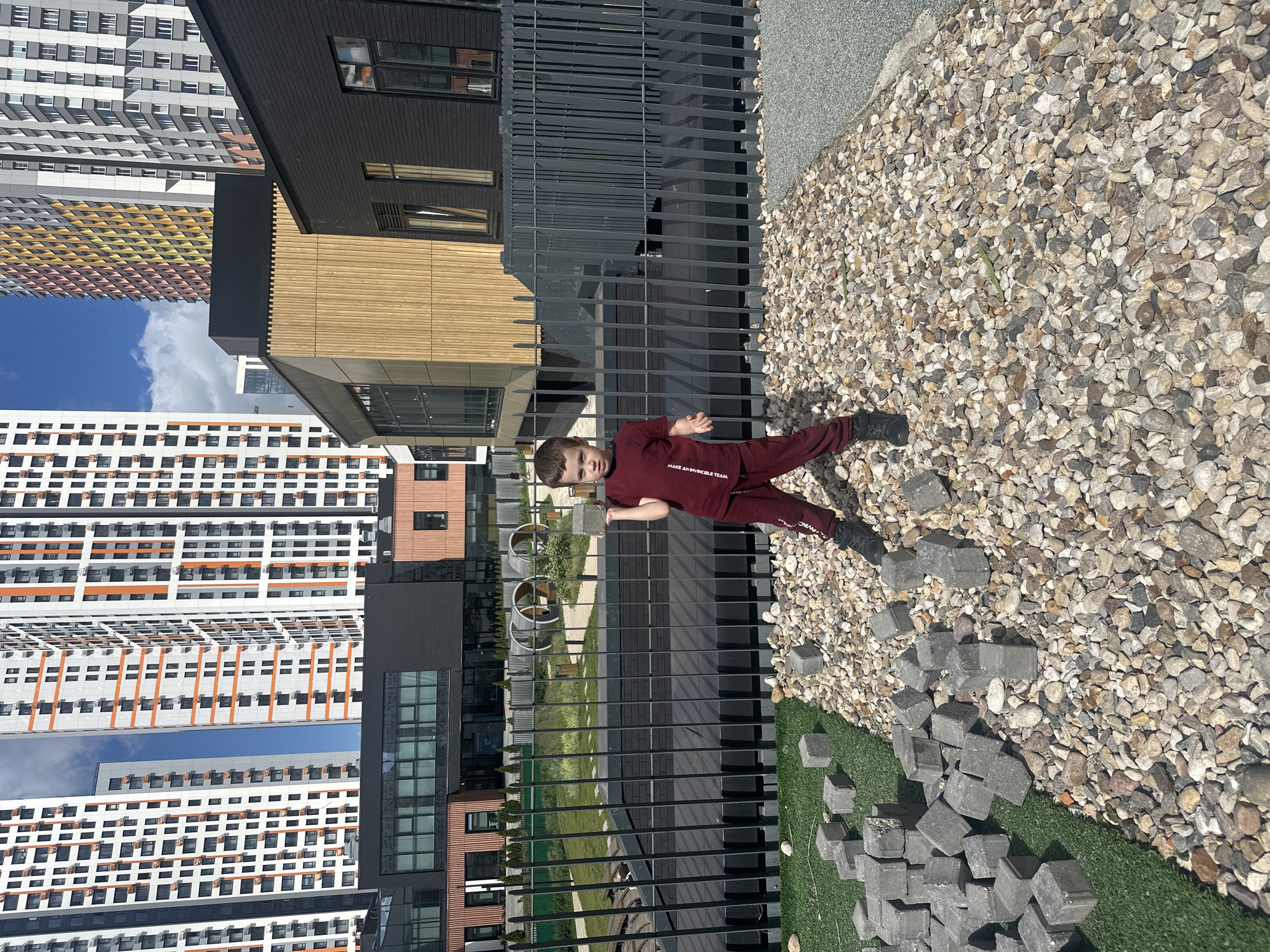 A young boy wearing a maroon t-shirt and matching pants stands on a gravelly surface, holding a gray brick in his right hand. He is looking towards the camera. Behind him is a black metal fence and a modern building complex with a dark facade and wooden accents. In the background, several tall, colorful apartment buildings pierce the sky under a bright blue sky with scattered clouds. The ground in front of the boy is a mixture of gravel and green artificial turf with more gray bricks scattered on both surfaces.