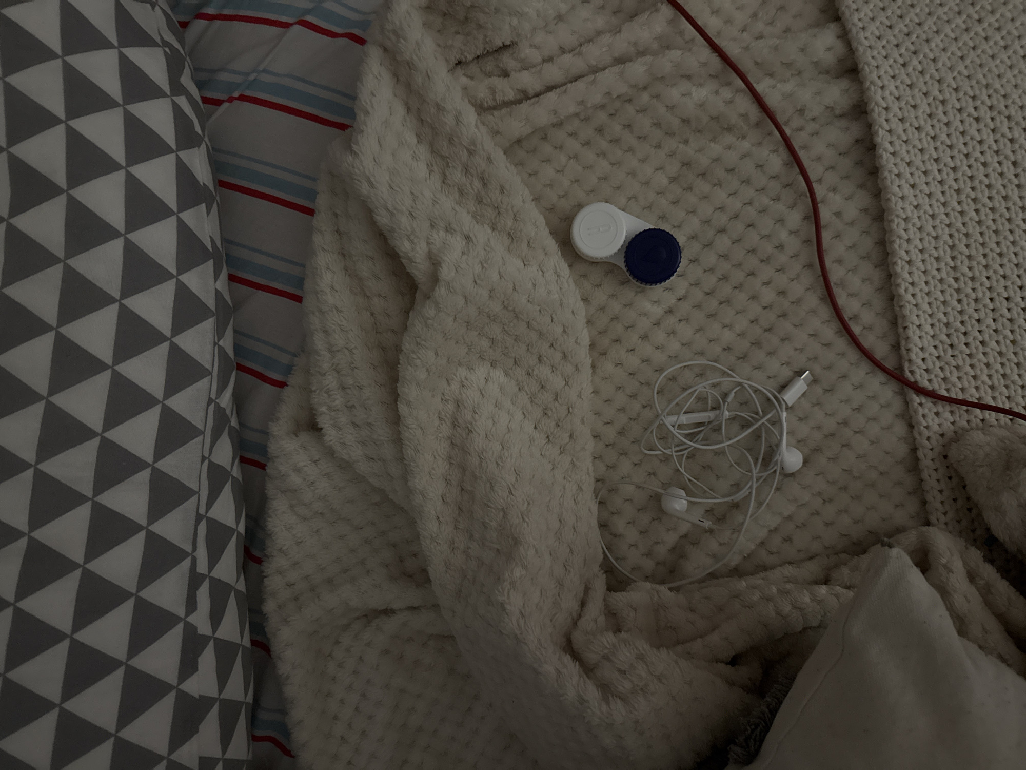 The image shows a close-up, overhead view of a bed with various objects. The bed is covered with a textured, cream-colored blanket. Partially visible beneath this blanket is a striped sheet with red and blue stripes. Above the blanket, a pillow with a geometric pattern of grey and white triangles is visible. On the blanket, a pair of white earbuds is tangled, and a small, white contact lens case with a blue lid is also present. A thin red cord is visible running diagonally across the lower right portion of the image. The scene appears to be an indoor setting, likely a bedroom. There is no indication of the city of Izobilnyy, Russia within the image.