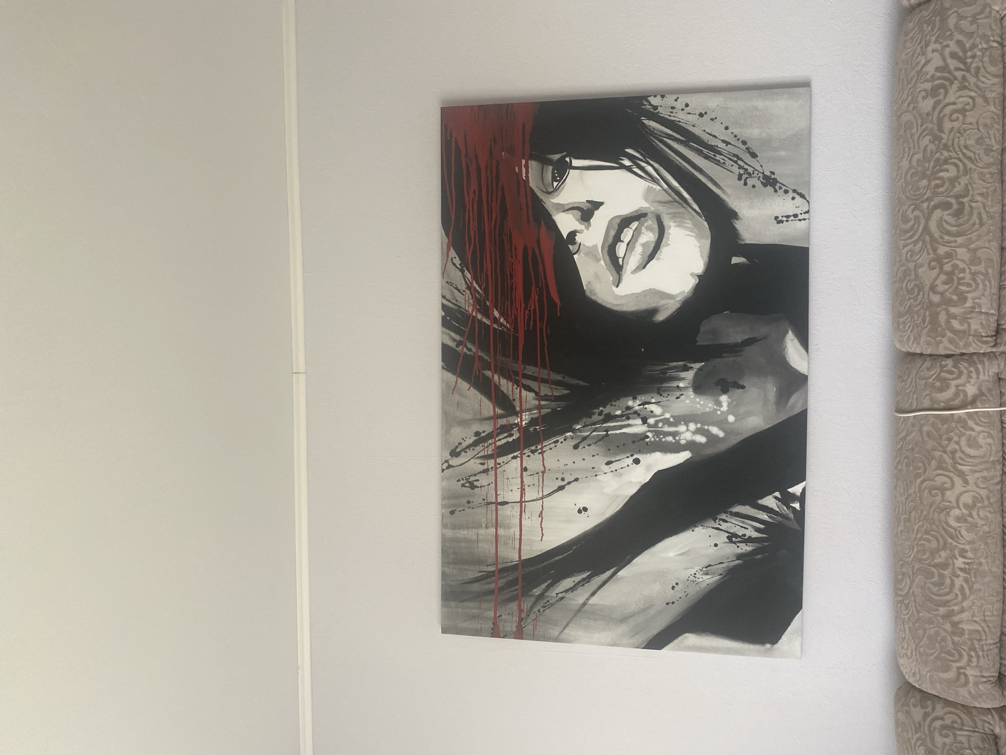 A large canvas painting of a woman's face, rendered in black and white with red accents, is hanging on a light-colored wall above a textured beige sofa. The woman's eyes are covered by horizontal red strokes, and her mouth is slightly open. The painting features a dynamic, abstract style with splatters and brushstrokes. The sofa has a patterned upholstery. The room appears to be a domestic setting, likely a living room. The image does not provide any specific information about the city of Krupki, Belarus, or any details relating to that location.