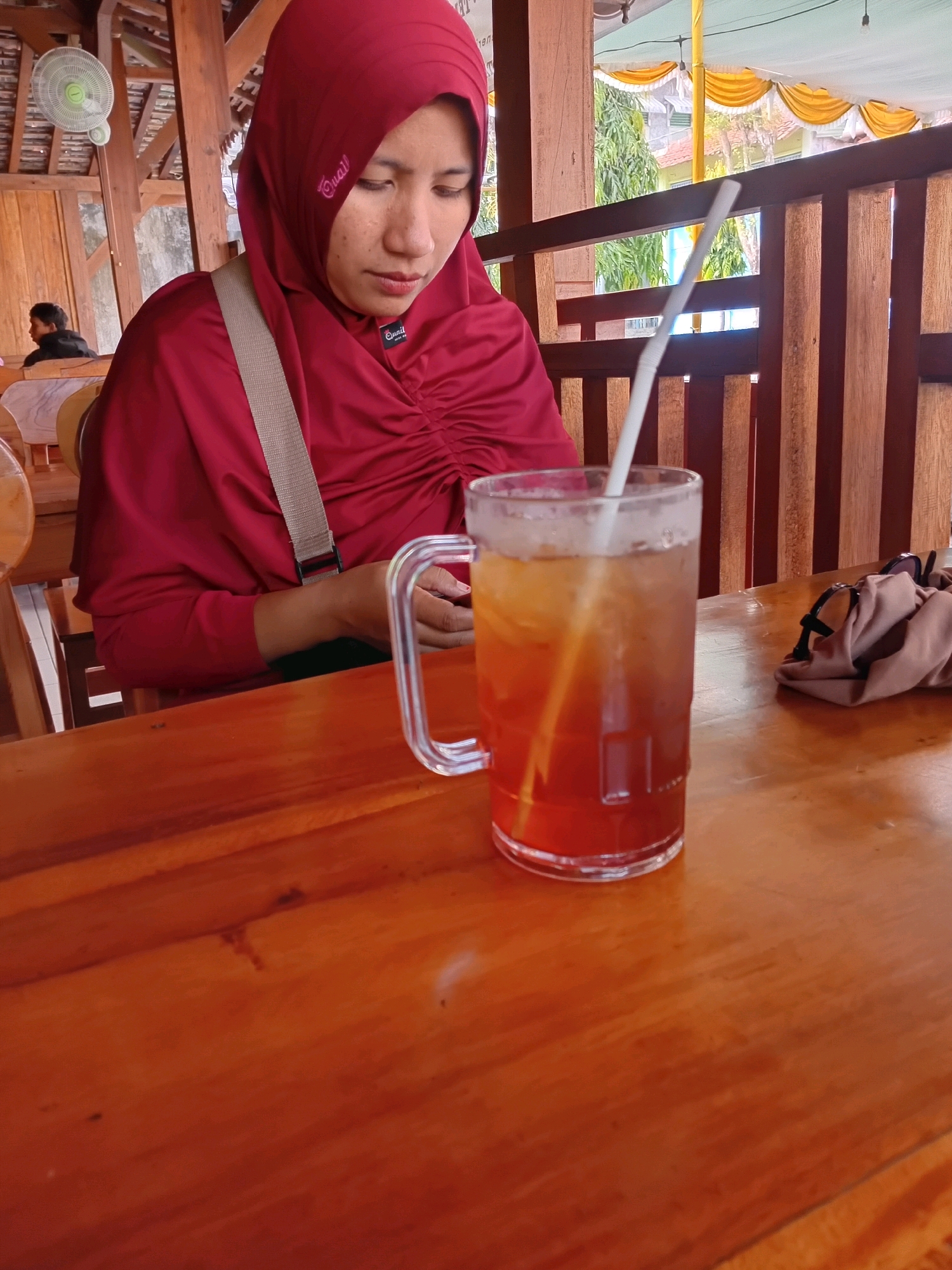 A woman wearing a red hijab and a red shirt is seated at a wooden table, with a glass of iced tea and a straw in front of her. The table is covered with a dark wooden finish. In the background, a wooden partition separates the seating area from the outdoors. A second person, a young boy, is visible in the background, seated at another table. The environment appears to be an open-air cafe or restaurant, with a thatched roof structure and bamboo elements visible. Sunlight filters through the open spaces, illuminating the scene. The overall impression is of a casual dining setting in a tropical or sub-tropical climate. The specific location in Purworejo, Indonesia, is suggested by the presence of local elements in the architecture and greenery outside.
