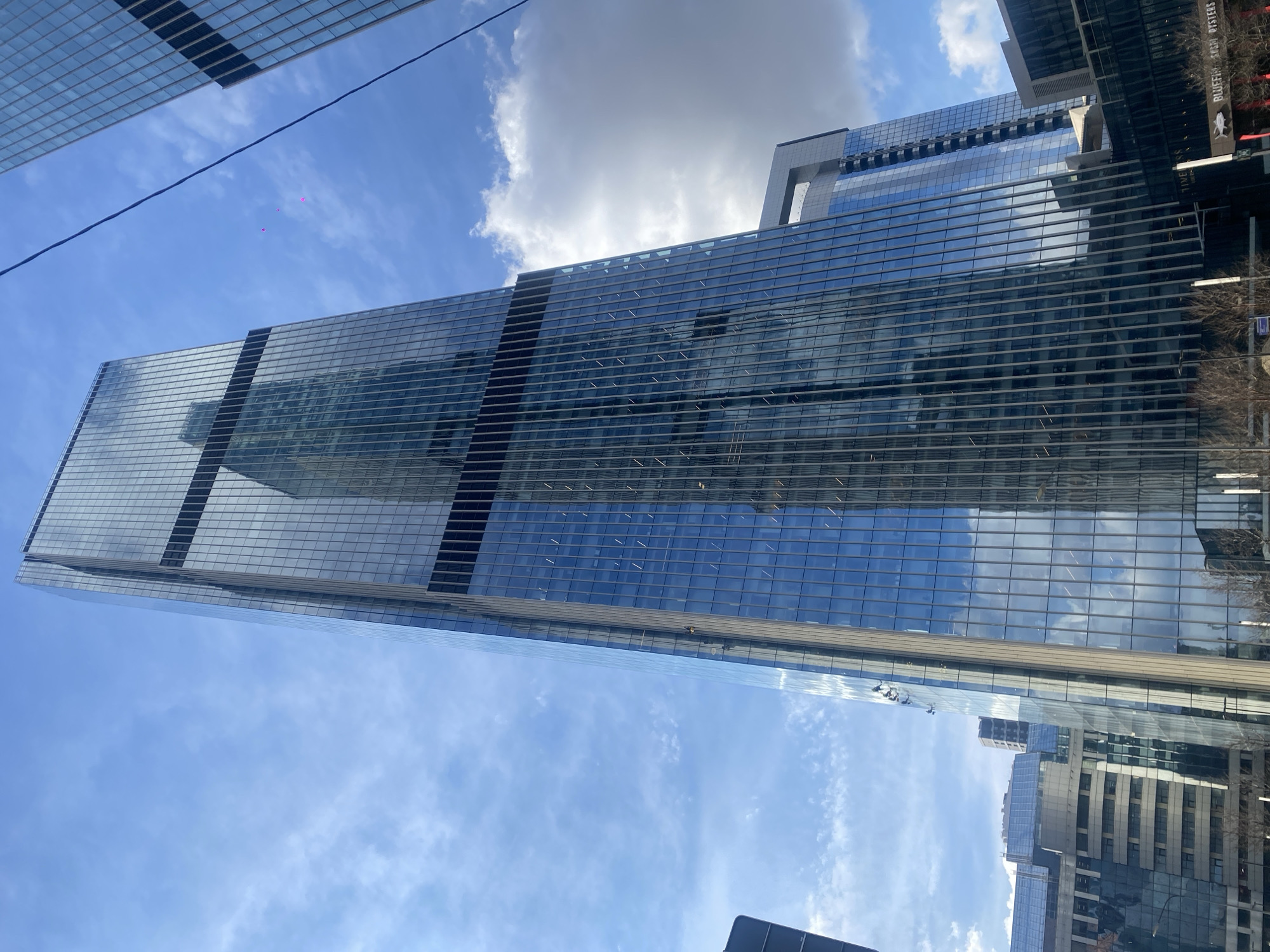 A low-angle, outdoor shot depicts a tall, glass-faced skyscraper against a partly cloudy blue sky. The building reflects the sky and other structures. Other buildings and trees are visible along the sides. A dark power line and three pink objects intersect the sky above the building. There is no evidence to support the location being in Khimki, Russia.
