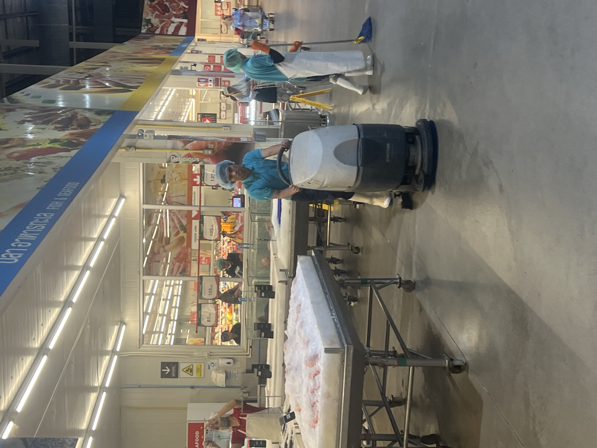 A bright, wide-angle interior shot captures a busy scene within a modern supermarket, likely a Makro store in Ko Samui District, Thailand, given the signage and context of the goods. The environment is a large, well-lit commercial space with a polished concrete floor. The ceiling features long rows of bright fluorescent lights.
In the foreground, a young man, wearing a turquoise short-sleeved shirt, white boots, and a blue hairnet, operates a floor scrubbing machine. The machine is grey and black, with "Nilfisk" branding visible. His posture suggests focus on the task.
To the right of the floor scrubber, another individual, dressed in a blue shirt, white apron, orange gloves, and white boots, is actively mopping the floor with a blue mop. They are also wearing a green hairnet.
In the mid-ground, several refrigerated display units are visible. One unit in the left foreground is filled with what appears to be fish or seafood on ice, covered in a white, foamy substance. Behind this, other display tanks contain live seafood. Further back, shelves display various goods, and posters with pricing, such as "85.00," "105.00," and "135.00," are visible. A woman in a white headscarf and dark clothing is seen behind a counter, possibly an employee. Another woman in a light-colored hijab and dark skirt stands near a shopping cart.
In the background, more aisles and displays are visible, including a large banner across the top of the store with Thai script and the words "FISH & SEAFOOD." This banner has a colorful mural of food items.
On the far left, a person in a red shirt and apron is seen from the shoulders up, holding a phone and seemingly taking a picture.
The overall impression is of a well-maintained, large-scale retail environment where food products, particularly seafood, are prominently displayed, and staff are actively engaged in cleaning and customer service.
