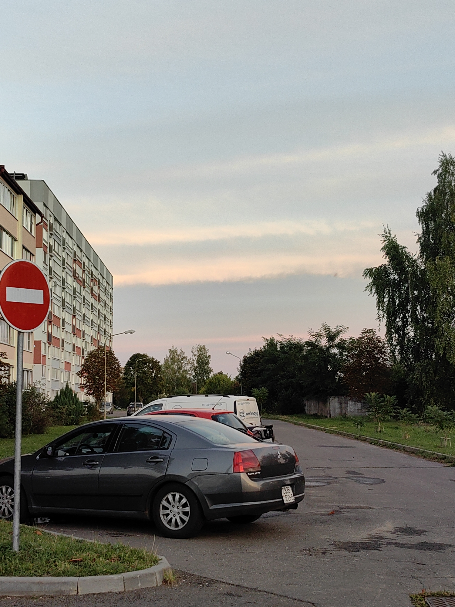 A grey sedan is parked on the right side of an asphalt road in front of a multistory apartment building on the left. The building has a facade of beige, red, and white rectangles. A red and white no-entry sign is planted on the left side of the road, in front of the building. The sky is a pale blue with soft pink clouds. Behind the sedan, a white van is parked. Trees and a low concrete wall are visible in the background.