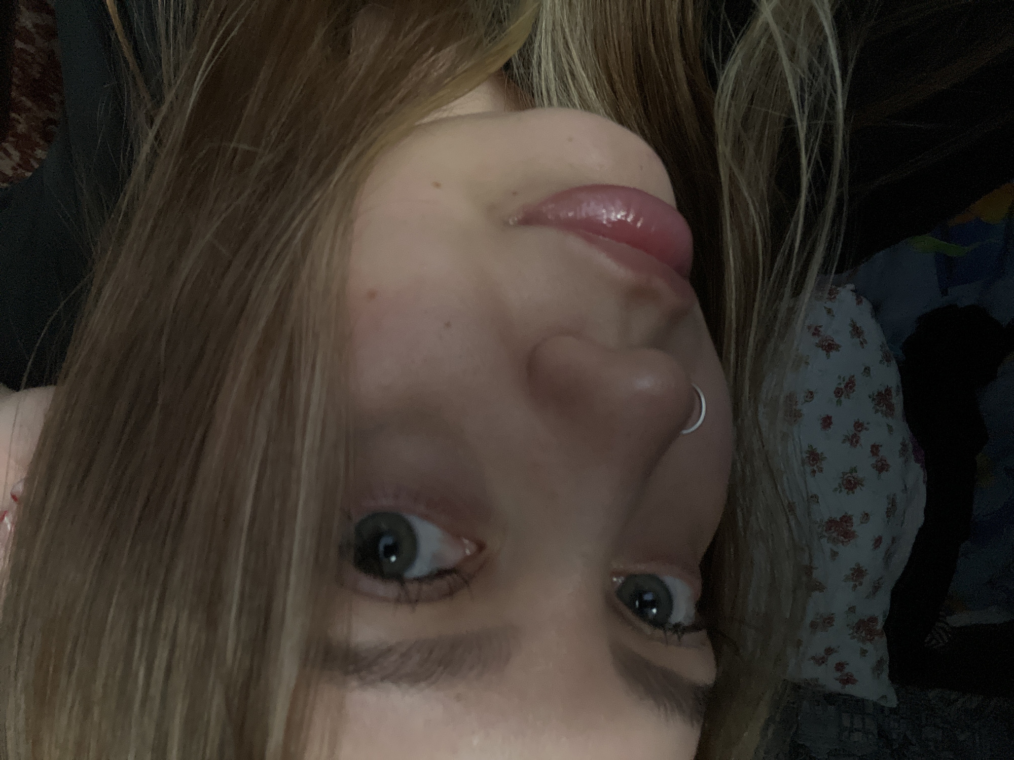 The image is a close-up, upside-down selfie of a young person with blonde hair. The subject's face is the primary focus. Their eyes are open and looking towards the camera. They have a small silver hoop piercing in their left nostril. Their lips are slightly parted, and their tongue is visible, appearing to be licking their lips. The lighting suggests an indoor setting, possibly in the evening or with artificial light. The background is blurred, but hints of fabric with a floral pattern are visible, as is some dark clothing. The overall mood is playful and intimate due to the close-up nature of the shot and the subject's expression. There is no visible text in the image.