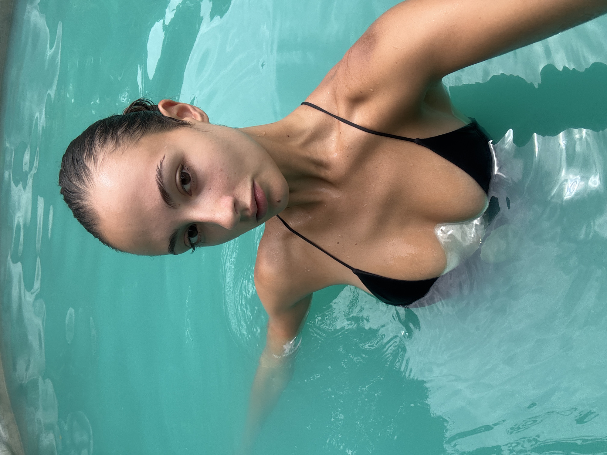 A woman is shown from an overhead angle, submerged in a turquoise-colored body of water, likely a pool or hot tub. She is wearing a black bikini top. Her dark hair is slicked back. Her left arm is extended into the water and her right arm is out of view. The location is potentially Ban Khlong Khian, Thailand.