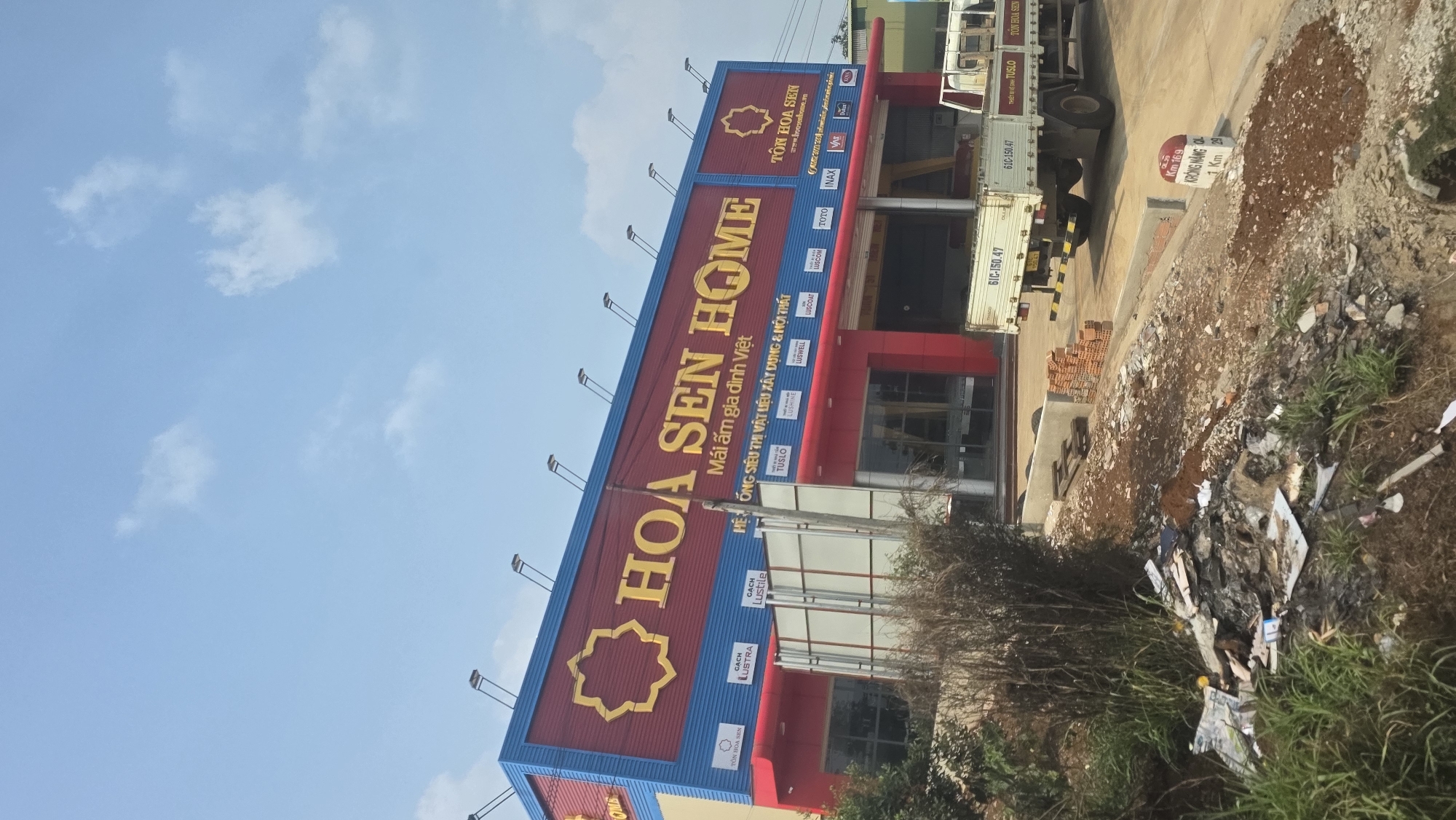 The image shows a commercial building in Trung Hồ, Vietnam. It is a "Hoa Sen Home" store, a chain specializing in building materials and home furnishings. The building is painted with a blue and red color scheme and is decorated with advertisements for various brands. Debris and vegetation can be seen in the foreground. Several vehicles can be seen. There is a kilometer marker beside a road reading Km 169 KRÔNG NĂNG QL.