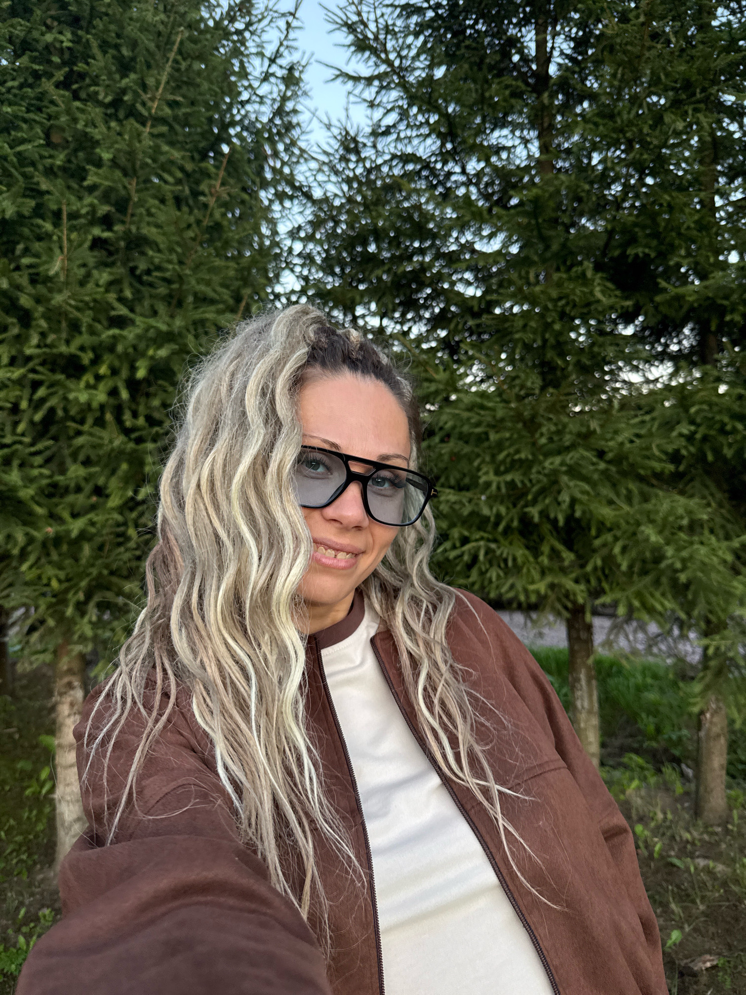 In Rusanovo, Russia, a woman with blonde, wavy hair and glasses takes a selfie outdoors. She is wearing a brown jacket over a white shirt. She is standing in front of a row of tall, green trees with a light sky visible through the branches. The ground is covered with grass and some foliage.