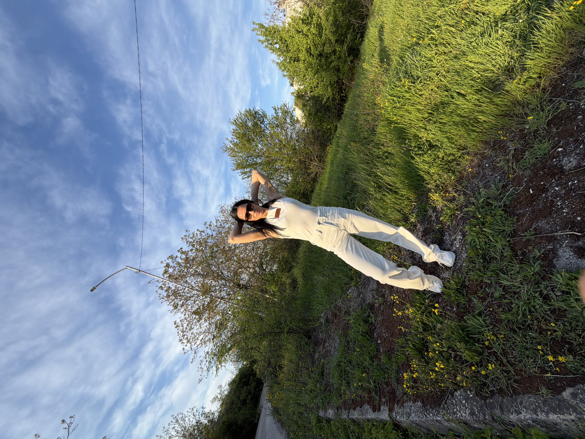 A full-body outdoor shot depicts a woman posing on a grassy slope under a bright blue sky with scattered wispy clouds, likely in Pleven, Bulgaria. The woman has long dark hair and is wearing sunglasses, a light-colored (possibly white or off-white) jumpsuit or overalls, and white sneakers. She stands with her arms raised, hands clasped behind her head, and looks directly at the camera, conveying a relaxed and confident demeanor.
The setting features a vibrant green hillside covered in grass and dotted with numerous small yellow flowers. Behind the woman and extending across the midground are several trees with rich green foliage. In the upper left, a tall, slender metal light pole with a single lamp reaches into the sky, with a thin wire stretching horizontally. A paved road or path is visible along the lower left edge of the frame, curving away. A low concrete or stone retaining wall separates the grassy area from what appears to be a lower embankment.
The warm, directional light and visible shadows suggest the photograph was taken during the late afternoon or early evening, likely the golden hour. The overall mood is bright and pleasant. There is no visible text in the image.