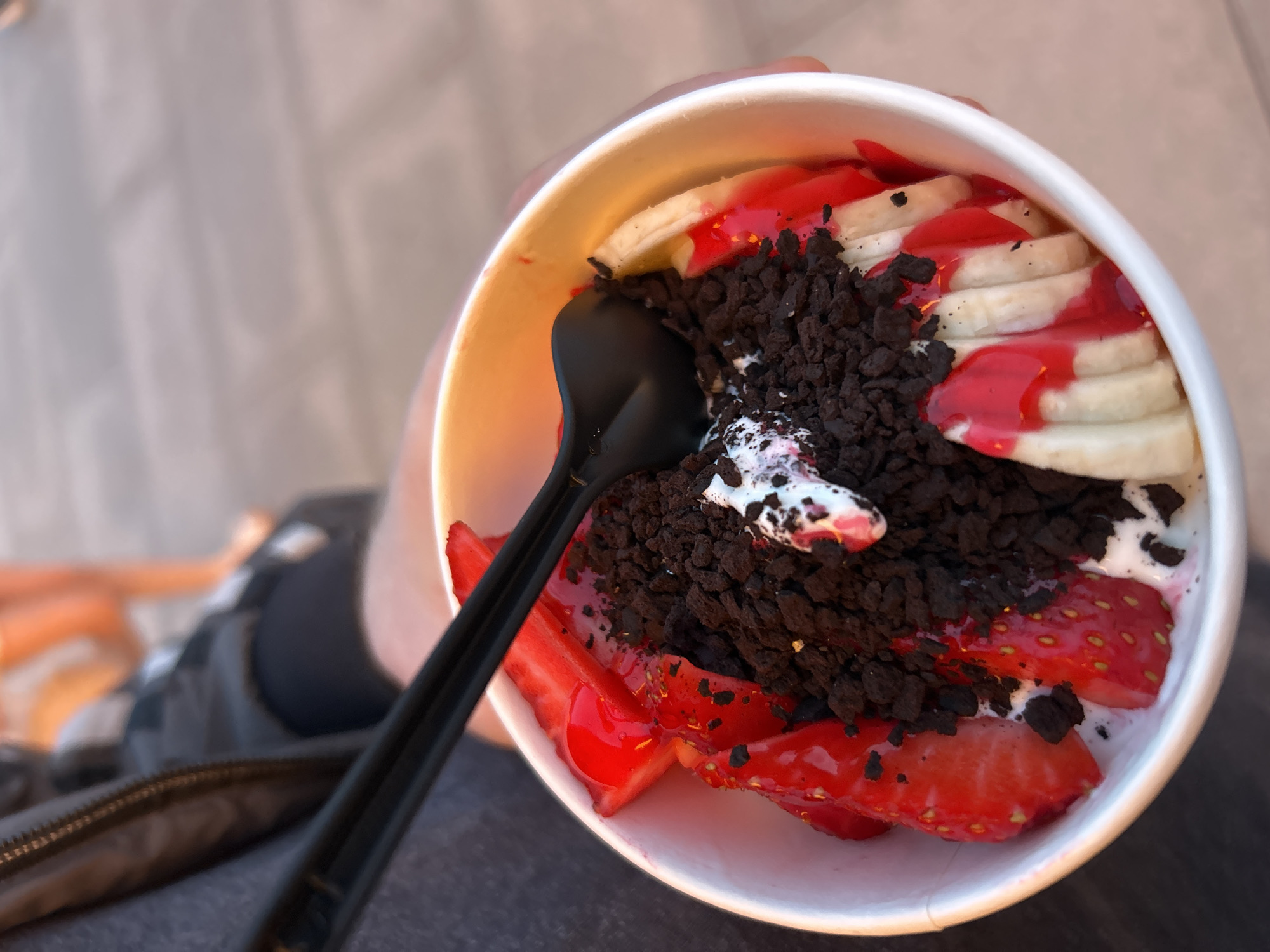 A close-up shot, taken from a slightly elevated angle, captures a white paper cup filled with a colorful dessert, likely frozen yogurt or ice cream, held by an unseen person. The dessert is generously topped with various ingredients.
Sliced bananas are arranged along one side of the cup, partially covered by a bright red, glossy syrup or sauce. Opposite the bananas, several bright red, sliced strawberries are visible, also drizzled with the same red sauce. The center of the dessert is piled high with dark brown, crumbled chocolate cookies, creating a rich, textured contrast. Hints of white cream or frozen yogurt peek through the toppings. A black plastic spoon is positioned vertically in the center of the cup, amidst the chocolate crumbles.
The hand holding the cup is only partially visible on the left side of the frame, blending into the blurry background. The background itself is out of focus, showing indistinct shapes and muted colors, suggesting a person's clothing (dark fabric, possibly a zipper from a bag or jacket) and a light-colored surface further back.
No specific time of day, weather conditions, or identifiable location cues are present. There is no visible text in the image. The focus is entirely on the appealing dessert, showcasing its vibrant colors and various textures.