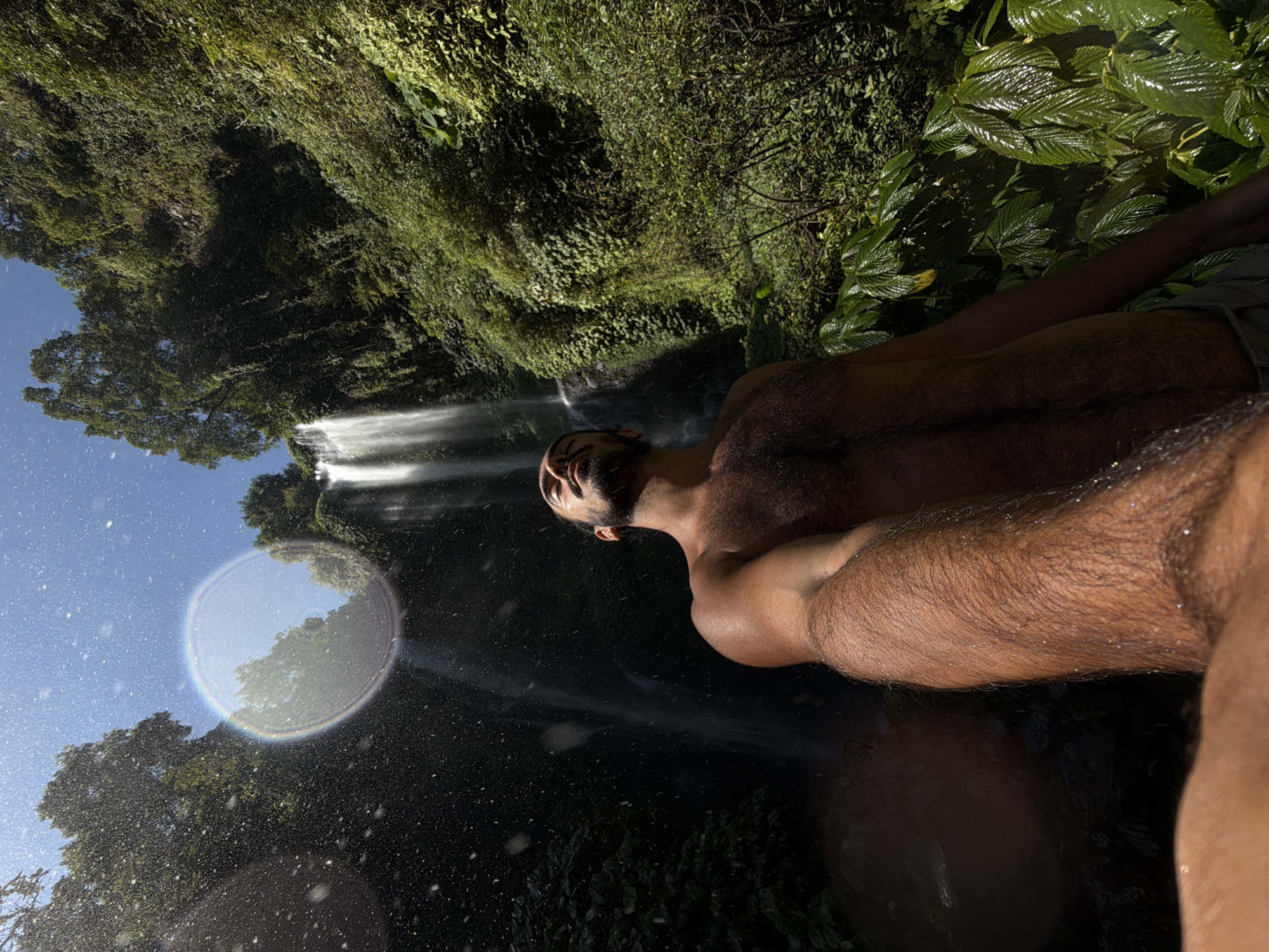 A low-angle, wide shot captures a man standing in front of a waterfall in a lush, green jungle environment. The man, shirtless and with dark hair, is looking up towards the sky with his eyes closed, seemingly enjoying the cascading water and mist. The waterfall is tall and narrow, with white water flowing down a rocky cliff face. The surrounding jungle is dense with vegetation, including large green leaves in the foreground. Sunlight filters through the trees, creating a bright, almost ethereal atmosphere. A circular lens flare is visible in the upper left quadrant of the image. The man's arm is extended towards the camera, and water droplets are visible on his skin and in the air. The scene suggests a natural, remote location, possibly a tourist attraction or a secluded spot for recreation in the Sawan region of Indonesia.