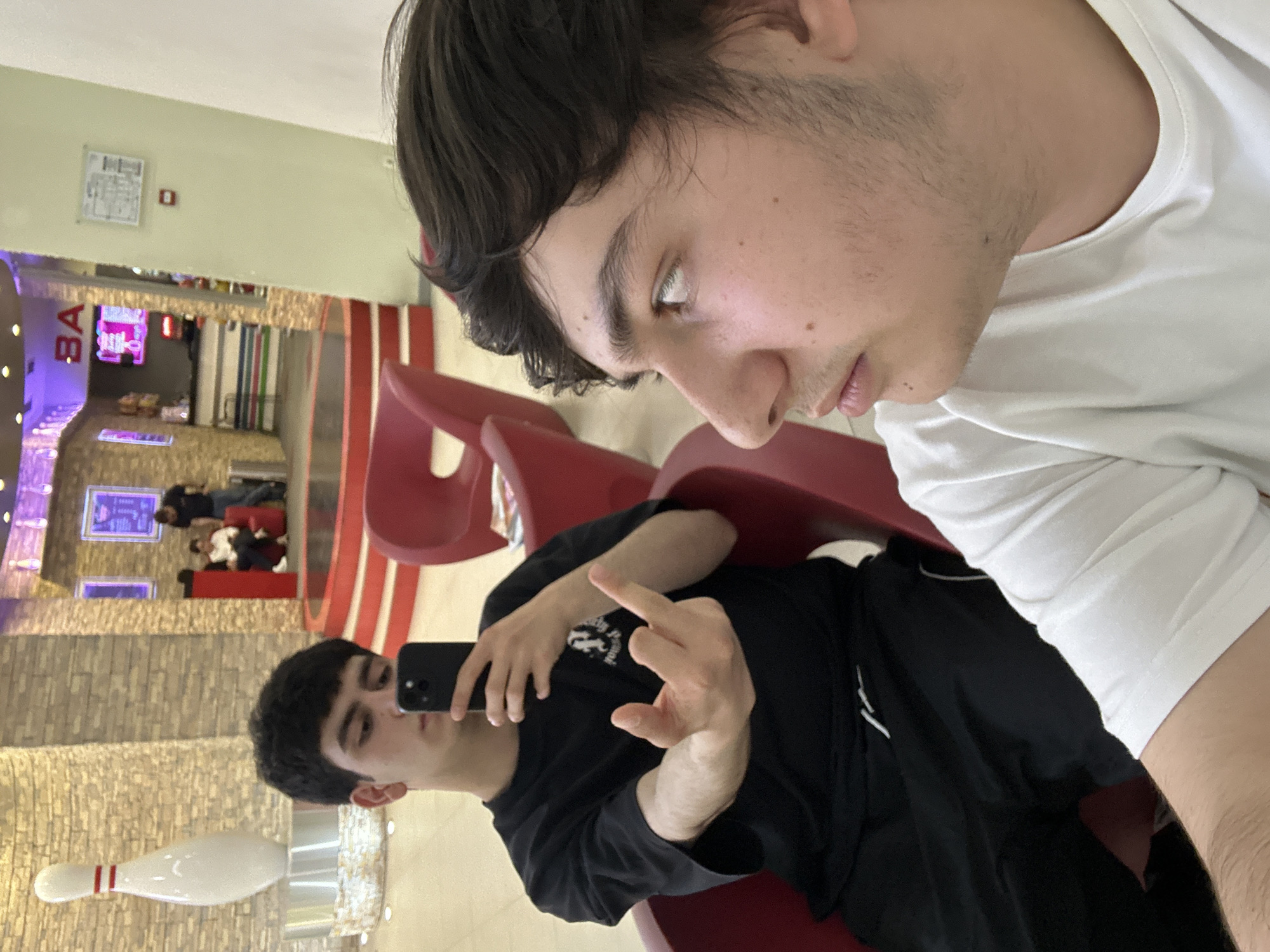 The image shows two young men sitting on a red bench in a bowling alley. The man in the foreground is wearing a white T-shirt and is looking down. Behind him, another man is wearing a black T-shirt and holding up his middle finger while taking a selfie with his phone. In the background, there are bowling lanes, a bar area, and several people. A bowling pin is visible near the bottom of the frame.