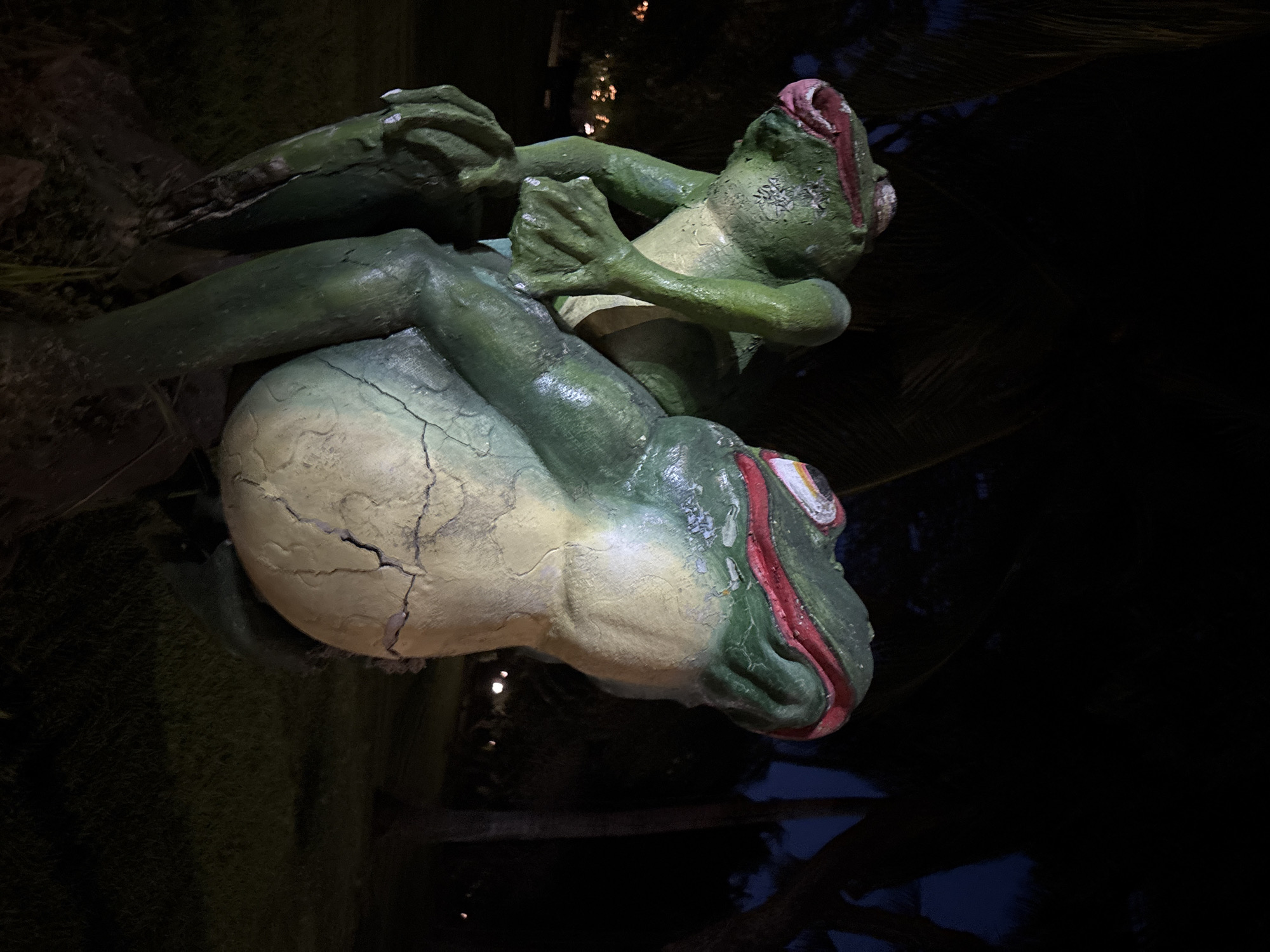 The image depicts a statue of two green frogs in Kuta Selatan, Indonesia. The statue is positioned outdoors, appearing at night or in very dim lighting. The lower frog is larger and cracked, supporting a smaller frog on its back. The top frog is also green, appearing to be kneeling. The background contains dark foliage.