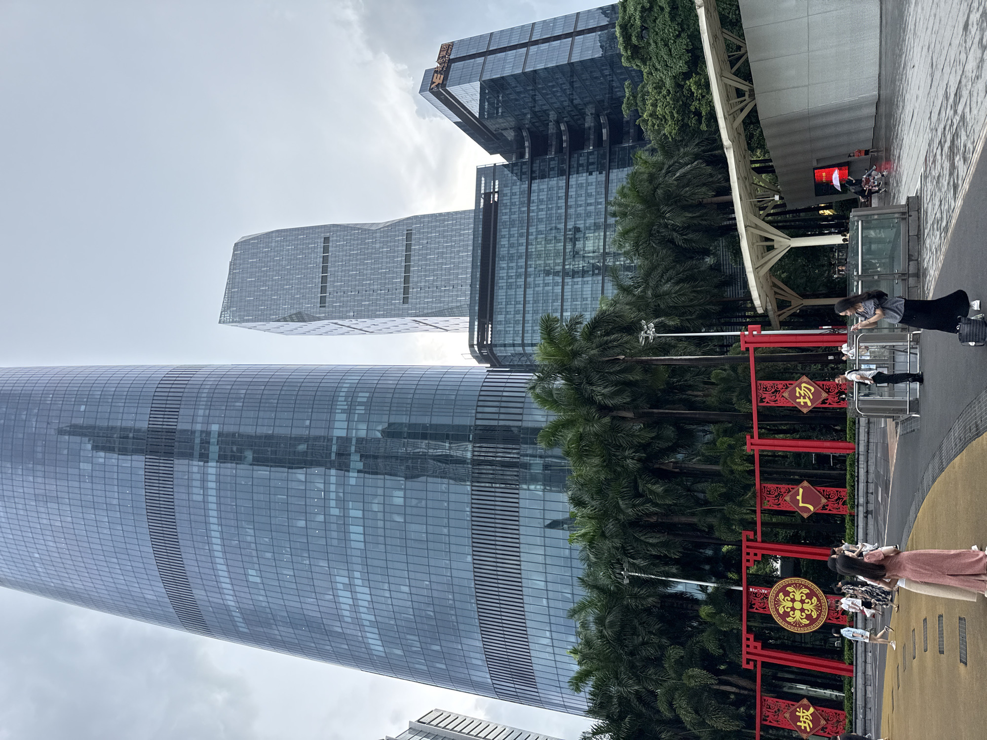 The image depicts a street scene in Tianhe District, China, characterized by modern skyscrapers, lush palm trees, and decorative banners. In the foreground, several people are visible. A woman in a striped pink and beige dress stands facing away from the camera, looking towards the right. To her left, another woman in a patterned dress is also looking away. Further to the left, a group of people, including a woman in a light blue dress and another in white, are walking. In the mid-ground, a red decorative archway spans across the path, adorned with traditional Chinese motifs and characters, including what appears to be "城" (chéng) and "场" (chǎng). Behind the archway, a dense line of palm trees stretches across the scene. Towering above the trees are several modern buildings, including a prominent, very tall glass skyscraper with a distinctive curved facade. To its right is another, somewhat shorter glass skyscraper with a more angular design. A third, darker glass building is positioned to the right of the angular skyscraper, featuring a logo that reads "Y.E. 永 集团". The sky is overcast, creating a diffused light. The ground is paved with a mix of yellow and grey material, with some puddles indicating recent rain. In the far right of the image, a white canopy structure extends over a pathway, and a large screen displays a red graphic. Additional people are seen near this structure, some holding umbrellas, and a child in an orange shirt.