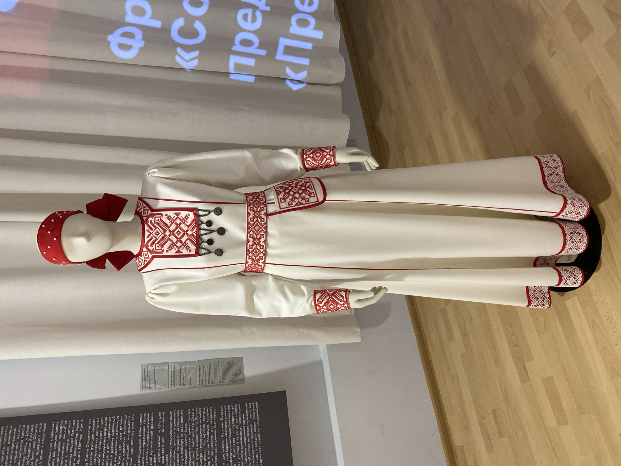 A white mannequin stands in a museum exhibit, wearing a traditional white dress with red embroidery. The dress features a red headscarf tied with a bow, embroidered cuffs, a wide embroidered belt with a small pouch, and a hemline with intricate red and white lace. The mannequin is positioned on a dark circular base, and the background consists of a light-colored wall and sheer curtains, with a projected image of blue text visible on the curtains. To the left of the mannequin, a museum information panel with text is mounted on the wall. The floor is made of light-colored wooden planks.
This exhibit is located in Petrozavodsk, Russia.