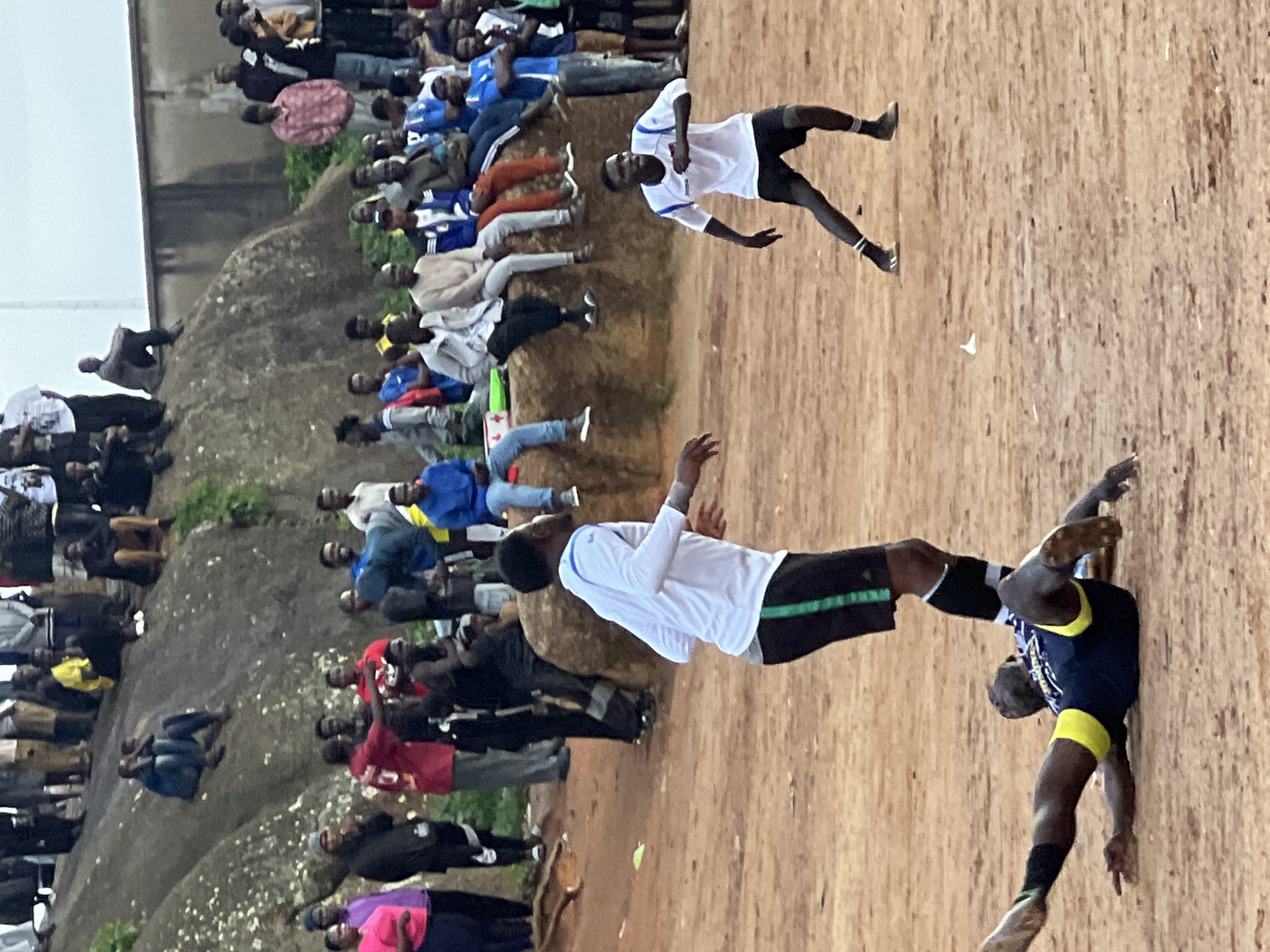 This image depicts a football match on a dusty, outdoor field in Jos, Nigeria. Spectators are visible in the background, some seated on rocky outcrops and others standing. The players are wearing football kits, with one player on the ground, seemingly having fallen during the game. The environment is characterized by dry, reddish-brown earth and sparse vegetation, typical of the landscape in Jos. The overall atmosphere suggests a local sporting event, with a mix of players and onlookers engaged in the activity.
