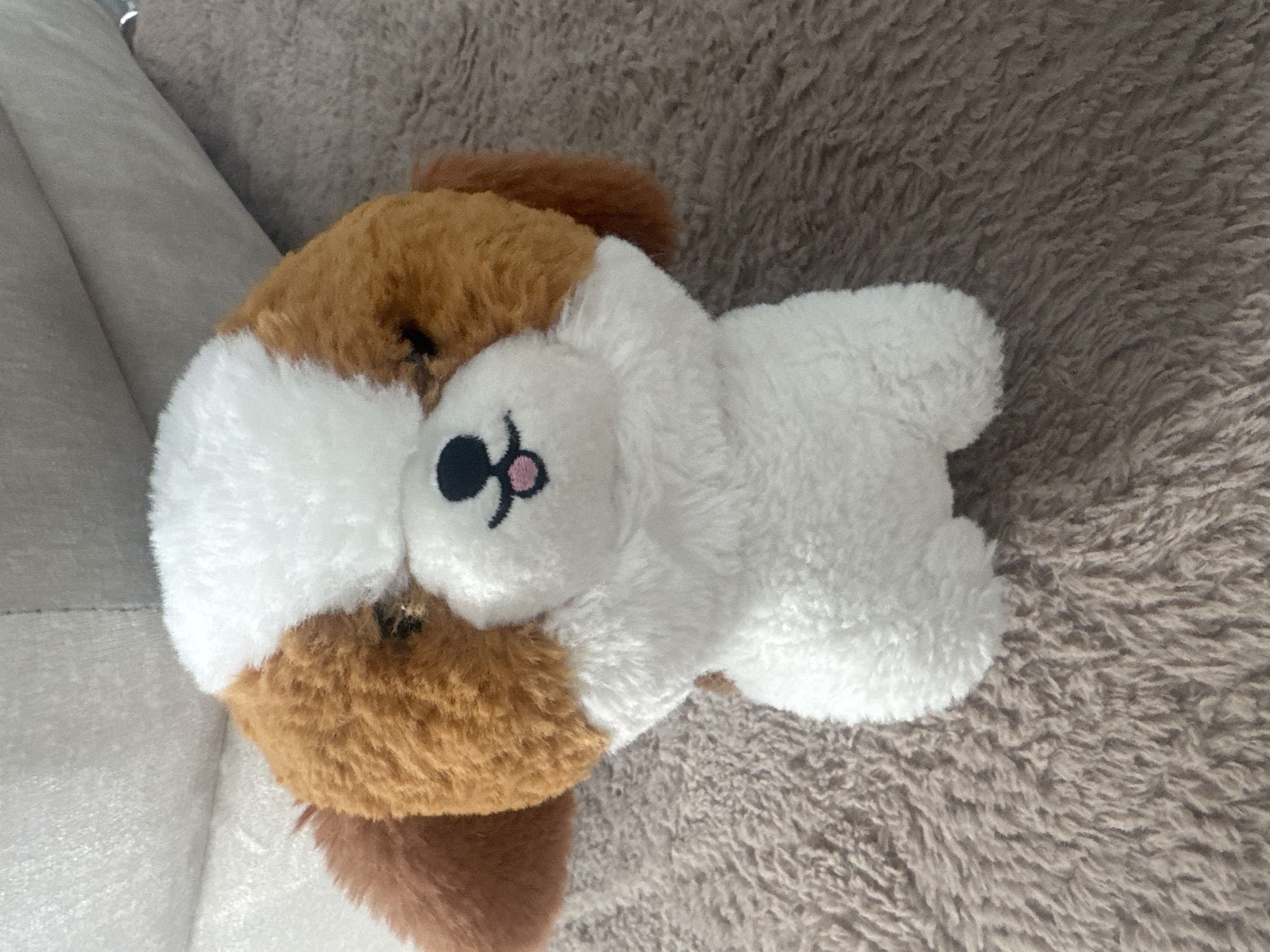 A stuffed animal, appearing to be a dog, lies on a brown, textured surface. The dog is mostly white with brown patches. Its face is white with a black nose and pink cheeks. Part of the dog rests against a gray object, possibly a sofa cushion. The surface underneath the dog appears to be a textured blanket or rug. The location is not discernable from the image.