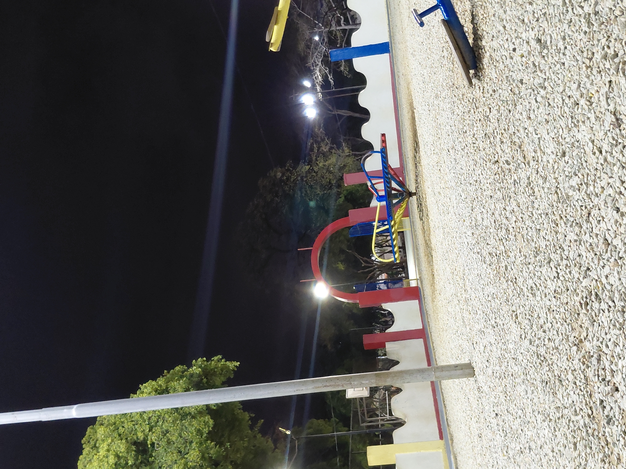 This image presents an unoccupied public playground at night in Santa Ana, Venezuela. The ground is covered with light-colored gravel. In the left foreground, a tall, light-grey pole stands before a tree with dense green foliage. A low, white wall with a curvilinear top edge, featuring sections painted red, yellow, and a blue vertical segment, extends across the midground. Behind this wall, a prominent red archway is visible. Playground equipment includes a multi-colored (yellow, blue, and red) spinning structure located behind the wall, and the blue end of a seesaw in the lower right foreground. Multiple artificial light sources illuminate the area, creating distinct lens flares against the dark, clear night sky. Further in the background, additional trees, some with bare branches, and a partial basketball hoop structure are present behind the wall on the left. No individuals or active interactions are observed.