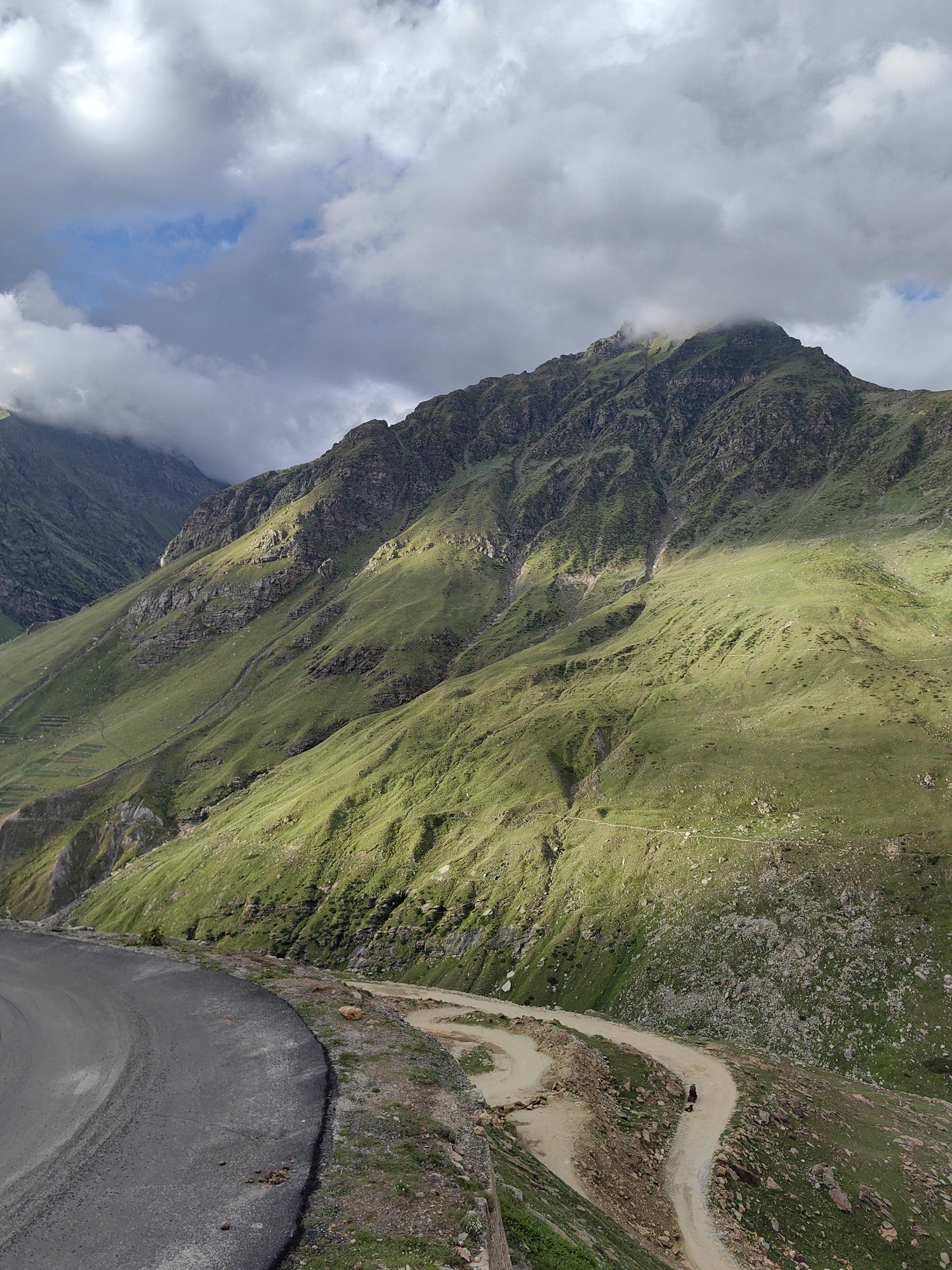 This is a photograph of a winding dirt road traversing a steep, grassy mountain slope in Gramphu, India. The road, characterized by sharp switchbacks, is the central focus of the lower portion of the image, with a section of paved road visible in the foreground. The mountain slopes are covered in vibrant green grass and dotted with rocks, indicating a high-altitude, rugged terrain. Sunlight casts dramatic shadows across the landscape, highlighting the contours of the mountains. The sky is a mixture of heavy gray clouds and patches of blue, suggesting dynamic weather conditions. In the distance, the peaks of the mountains disappear into the clouds. A single figure on a dark-colored motorcycle is visible on the dirt road, adding a sense of scale and activity to the otherwise natural scene. Small, terraced fields are subtly visible on a lower part of the mountain, hinting at human agricultural presence in the area.