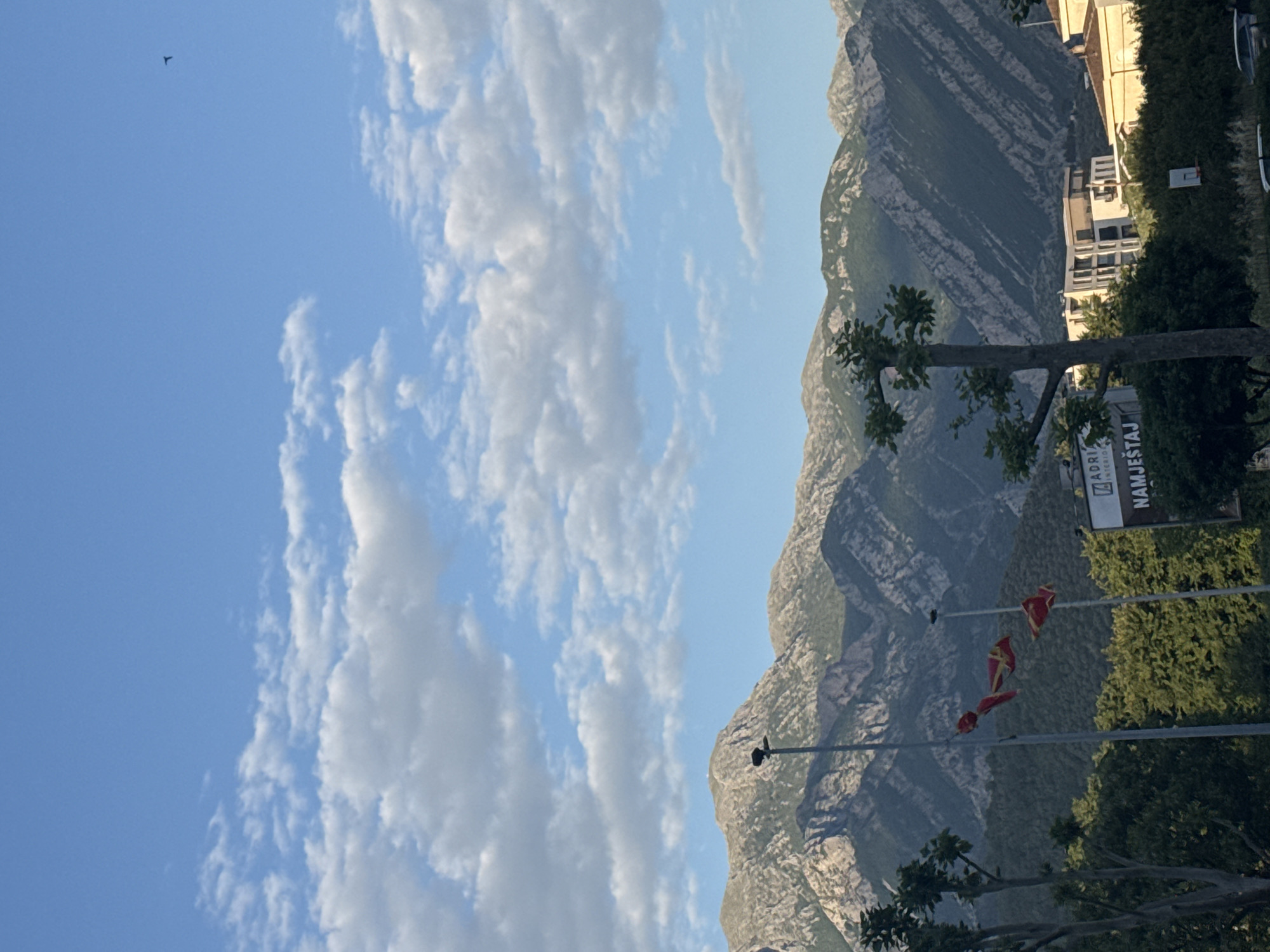 The image captures a scenic view of Bar, Montenegro. The foreground is a combination of trees and foliage, partially obscuring a sign that reads "ADRIA NAMJEŠTAJ." Three Montenegrin flags are hanging from wires stretched between utility poles. In the background, a towering mountain range dominates the landscape. Several buildings are situated on the mountainside. The sky is light blue, dotted with scattered clouds. In the upper-left corner, a small bird is visible in flight.
