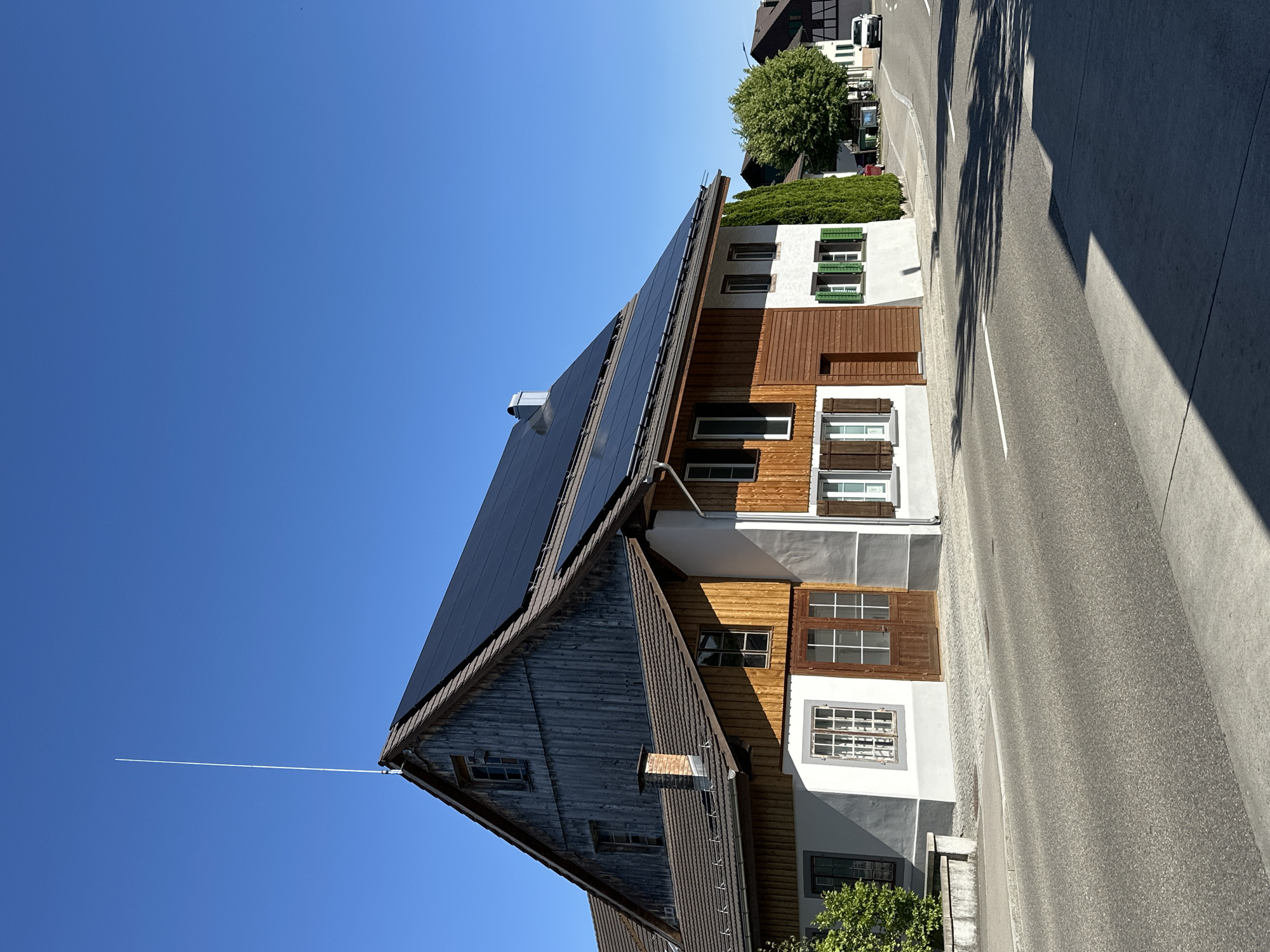 The image shows a residential building in Embrach, Switzerland, on a clear, sunny day. The building is a two-story structure with a dark-tiled roof covered in solar panels. The upper section of the building's facade is clad in weathered wooden planks, while the lower section is painted white with gray accents. It features several windows with white or green shutters and wooden double doors. A tall, thin antenna extends from the peak of the roof. The building is situated alongside a paved road with white lane markings. In the background, other buildings and trees are visible, and a white car is driving away from the camera. Shadows from trees and the building are cast across the road and sidewalk, indicating the position of the sun.