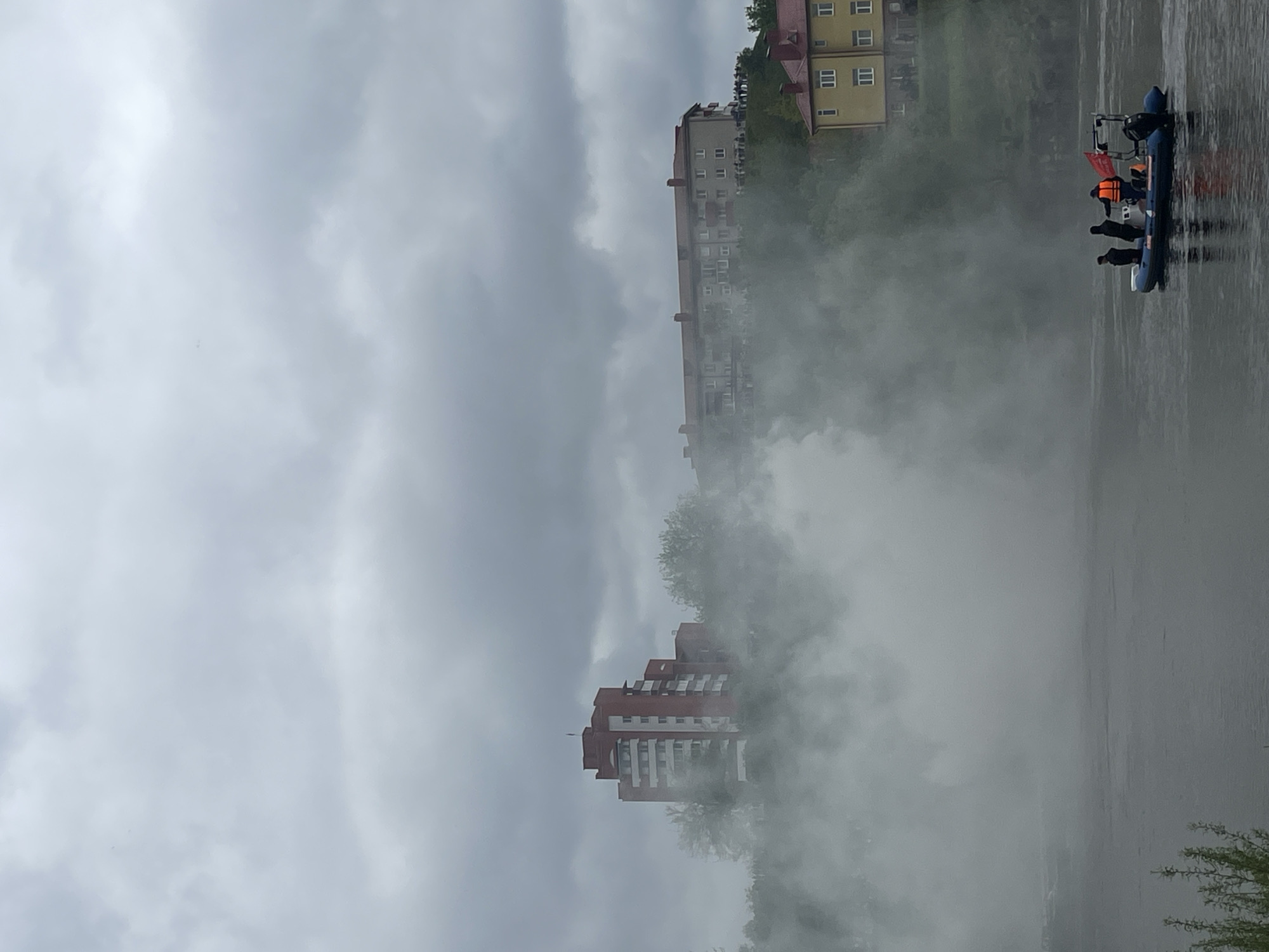 The image depicts a cityscape shrouded in mist, likely in Hrodna, Belarus, given the architectural style of the buildings. In the middle ground, several multi-story buildings with red and beige facades are partially obscured by the mist. To the right, a dark body of water reflects the sky. A small inflatable boat with several individuals aboard is visible on the water's surface. The individuals are wearing dark clothing, and at least one person is wearing an orange life vest. The sky is overcast, with heavy grey clouds dominating the upper portion of the image.