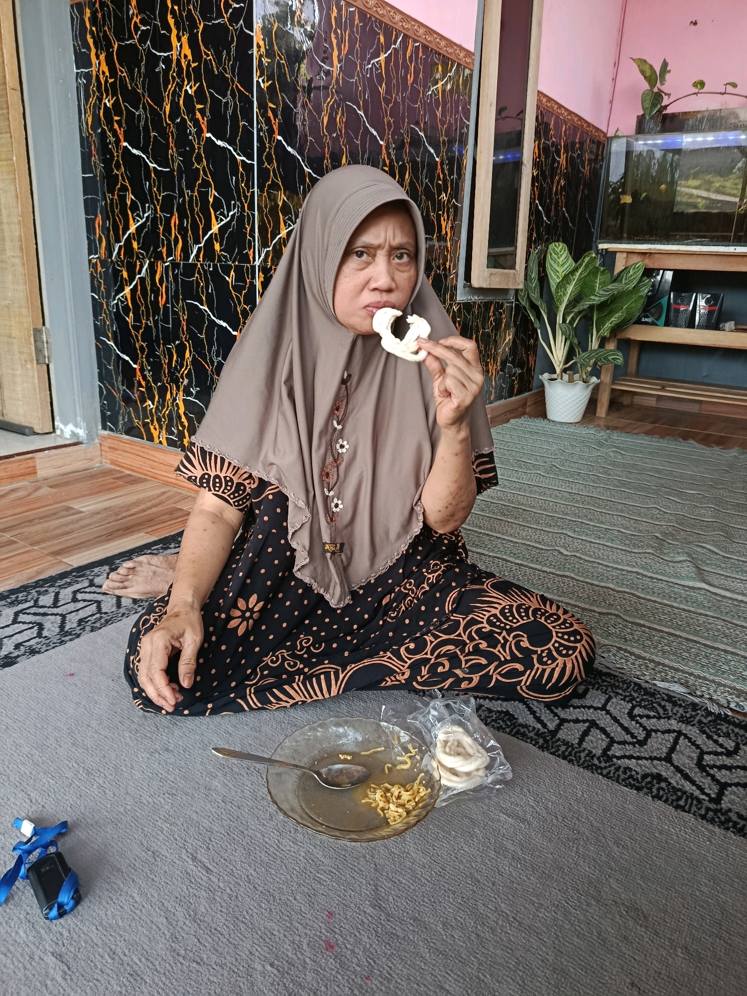 An adult Indonesian woman, wearing a brown hijab and a black and orange batik dress, sits on a gray rug in front of a wall with a black and orange marble-like pattern. She is holding a light-colored snack to her mouth with her right hand. In front of her, on the rug, is a small, clear plate with some noodles and a spoon, and a plastic bag containing more of the same snack. To the left of the plate, on the rug, is a small black object with a blue ribbon tied around it. In the background, to the right of the woman, is an aquarium filled with water and fish, and a green potted plant on a wooden shelf. The floor is tiled. The scene appears to be indoors, possibly in a home in Bululawang, Indonesia.