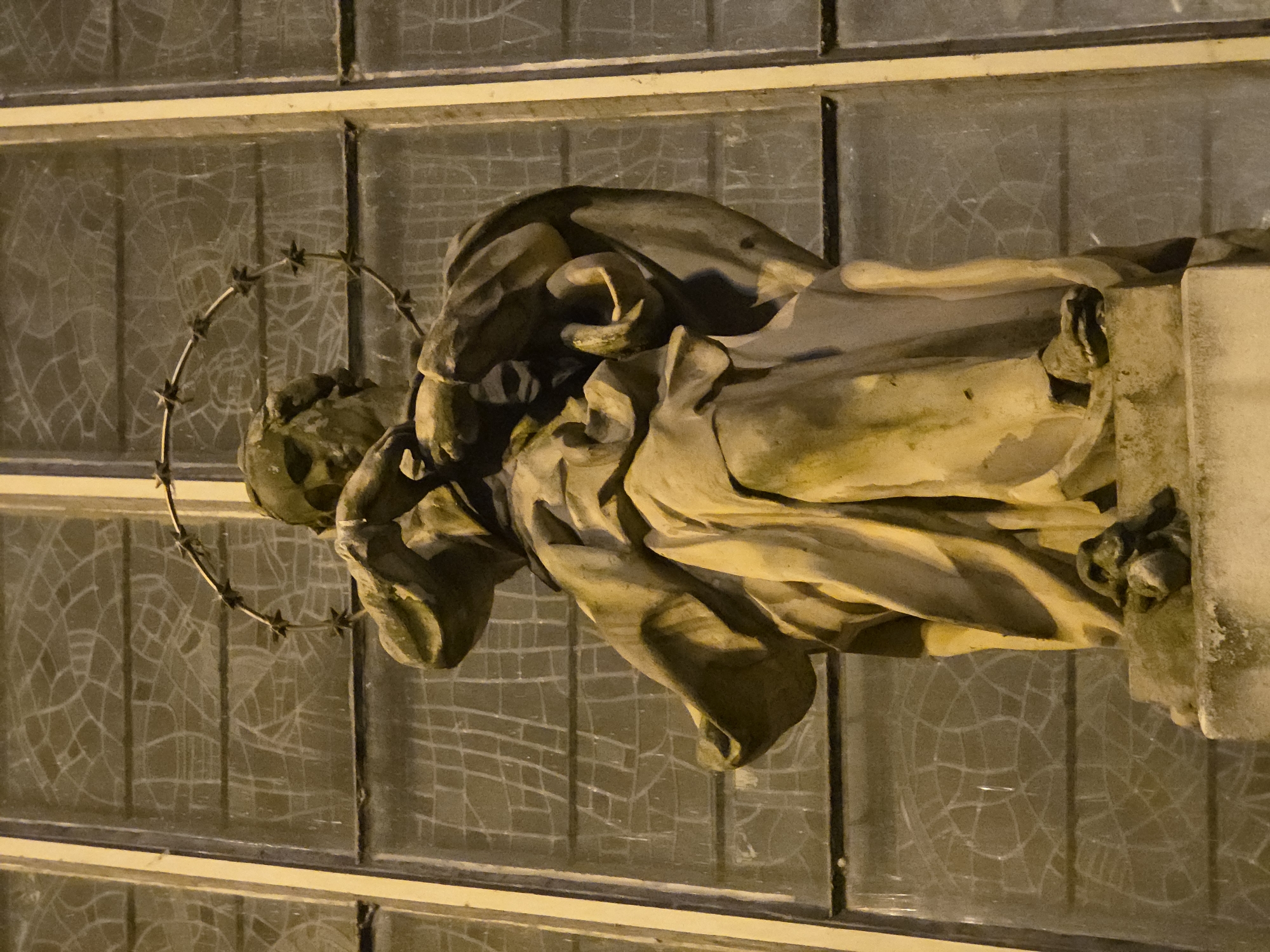 A nighttime close-up depicts a weathered statue of a religious figure, possibly Jesus, adorned with a barbed wire halo. The statue is positioned in front of a multi-pane window with a distinct cracked pattern in the glass. The statue is likely located in Warsaw, Poland, based on the broader context of the image set.