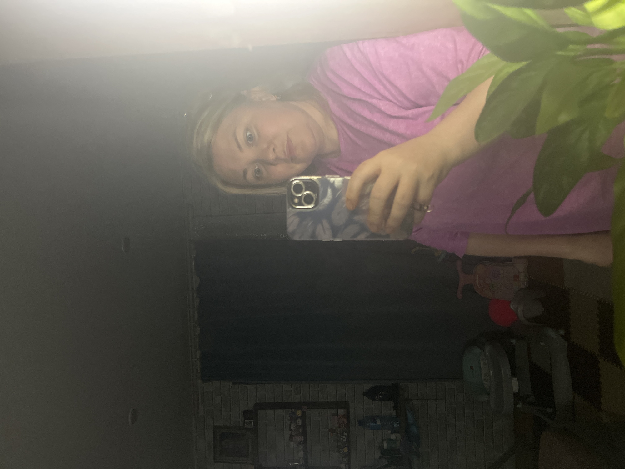 A woman with blond hair and blue eyes is taking a selfie in front of a mirror. She is wearing a pink shirt. The phone she is holding has a blue and white patterned case. Behind her is a brick wall with a collection of items on shelves. There is also a green plant to the right of the woman. The overall lighting is dim. The location is in Russia, but further information about it being Vesyoloye, Russia is not possible from the image.