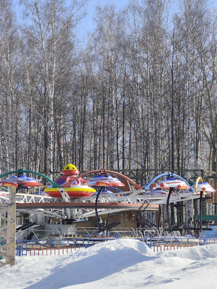 The image displays an amusement park ride situated in a snowy, wooded area of Chaikovsky, Russia. The foreground features a substantial accumulation of undisturbed white snow covering the ground. In the midground, a stationary amusement ride is visible, consisting of multiple colorful (red, yellow, blue, green) flying saucer-themed cabins mounted on a gray and white metal framework. The cabins are unoccupied, and the ride appears to be non-operational. A low, multi-colored barrier fence, featuring blue, orange, and white sections, encircles the base of the ride. The background consists of a dense forest of bare deciduous trees, predominantly birch, with light bark contrasting against darker branches. The sky above is clear and light blue. No individuals are present in the scene. The overall environment suggests a winter or early spring setting due to the snow and leafless trees.