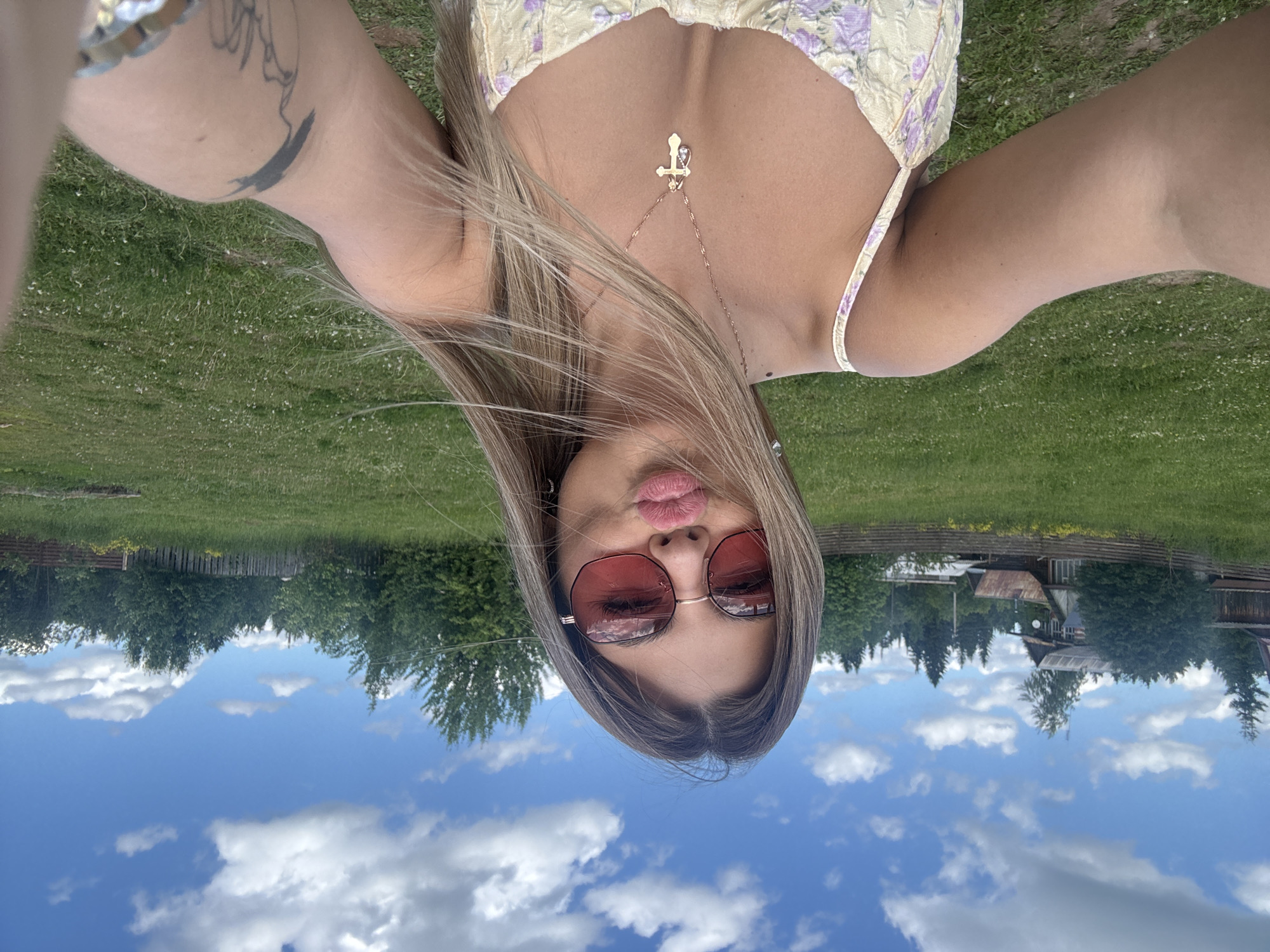 This media file shows a young woman taking a selfie outdoors. She is wearing a light yellow and purple floral crop top and sunglasses with pink lenses. A silver necklace with a cross pendant hangs around her neck. Her hair is long and blonde. She is holding her phone up to take the picture, and her lips are pursed.
The background suggests a rural setting in Pastukhovo, Russia. There is a grassy field with scattered wildflowers, a wooden fence, and trees. In the distance, there are some buildings, possibly houses or sheds, and more trees. The sky is blue with fluffy white clouds, indicating it is daytime and likely fair weather. The overall impression is casual and relaxed, with the subject appearing playful or posing. There is no visible text in the image.