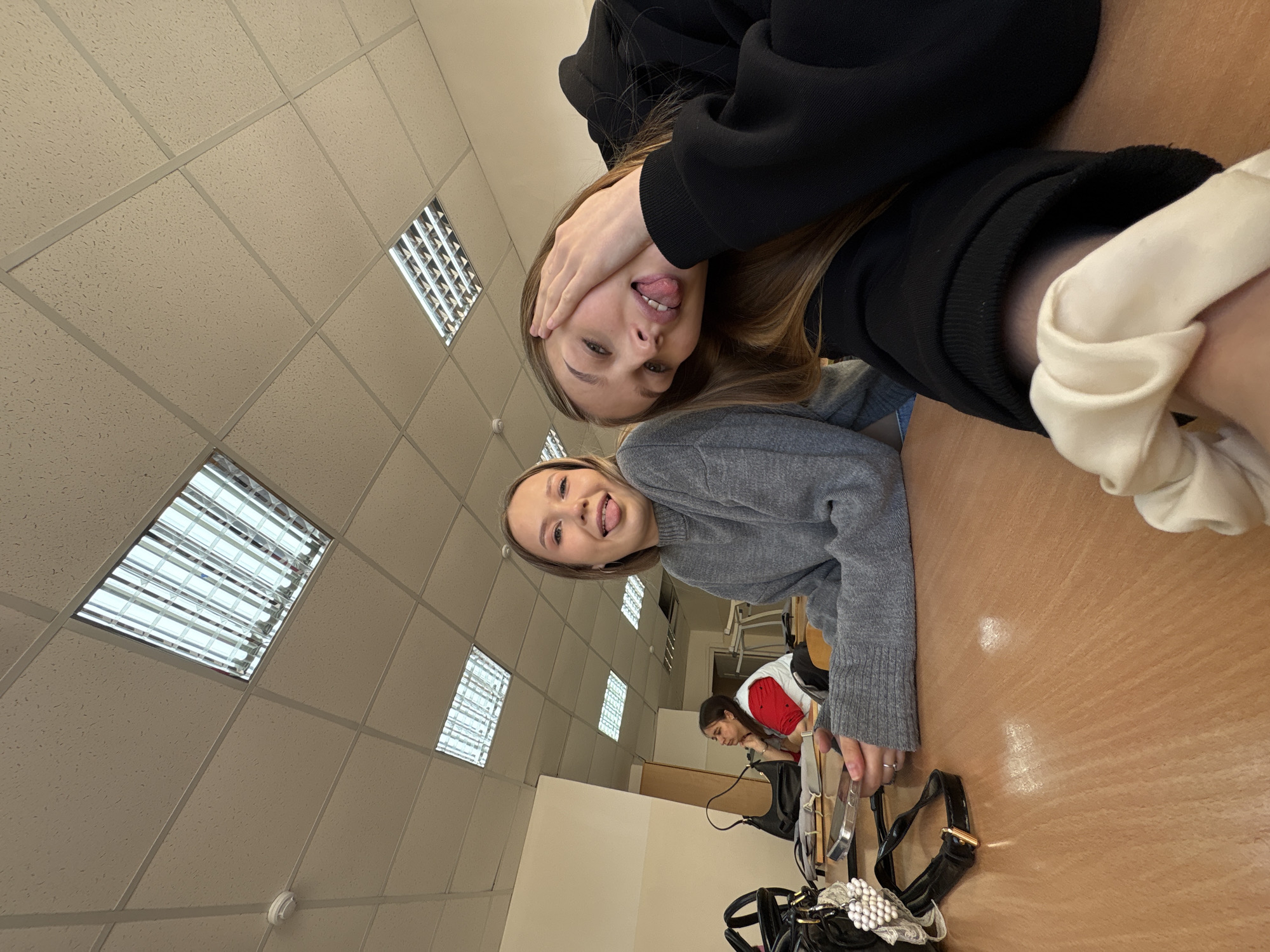 Two young women are posing for a selfie in what appears to be a classroom. The location is potentially in Chaikovsky, Russia, based on other metadata. The classroom has a drop ceiling with fluorescent lighting. One woman is wearing a gray sweater and the other a black hoodie. The woman in the hoodie is covering part of her face with her hand, sticking out her tongue. A desk is visible with handbags on it. In the background, another individual is working at a desk.