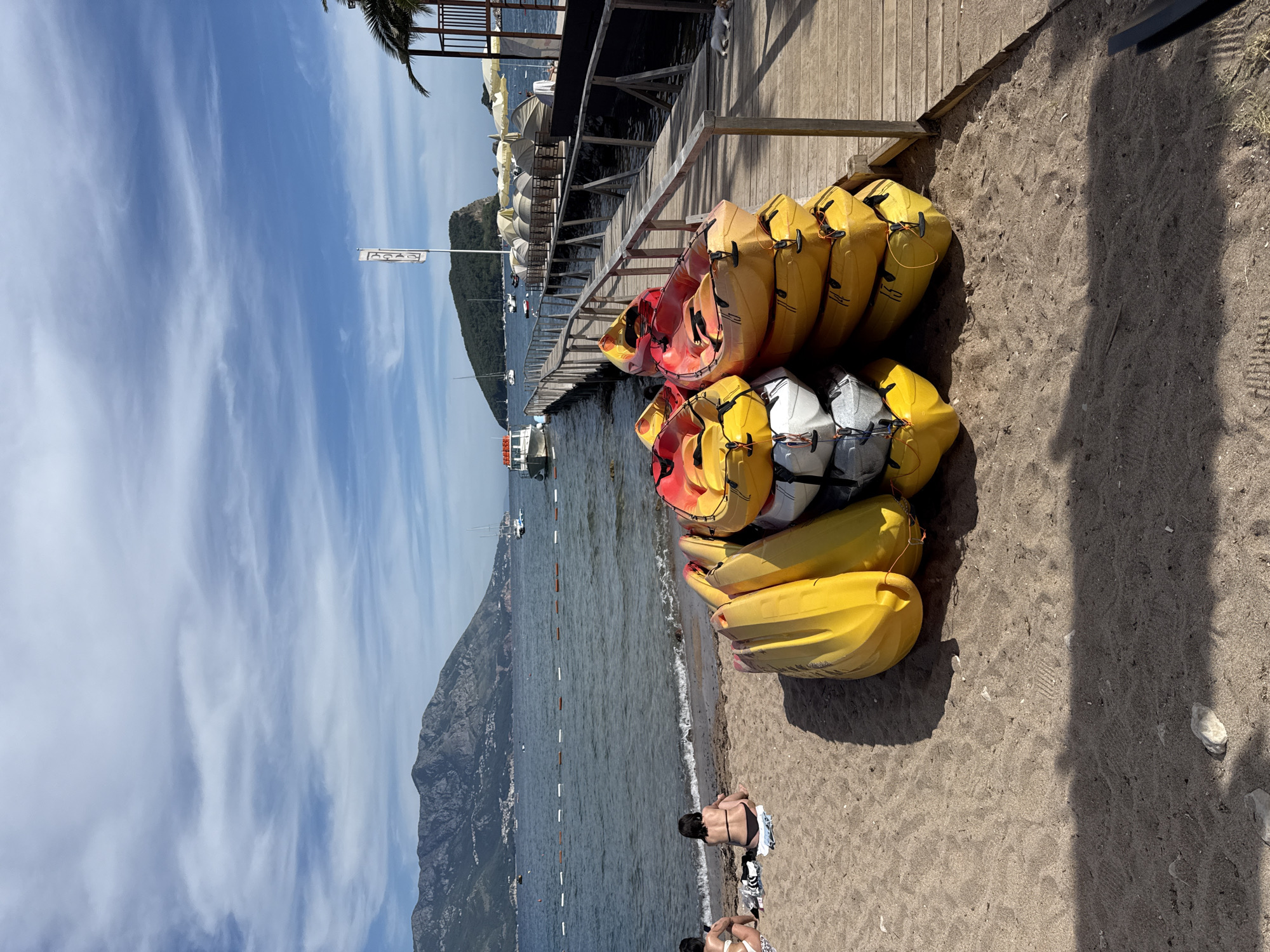A sunny day at a beach in Budva, Montenegro features a stack of yellow and red kayaks on the sand in the foreground. The kayaks are piled on top of each other, with some white kayaks also visible underneath. A wooden pier extends from the right side of the image into the sea, with lounge chairs and umbrellas set up on the pier in the background. Several boats are moored in the water, and mountains rise in the distance. Two women are sitting on the sand near the water's edge, facing away from the camera. The sky is blue with scattered clouds.
