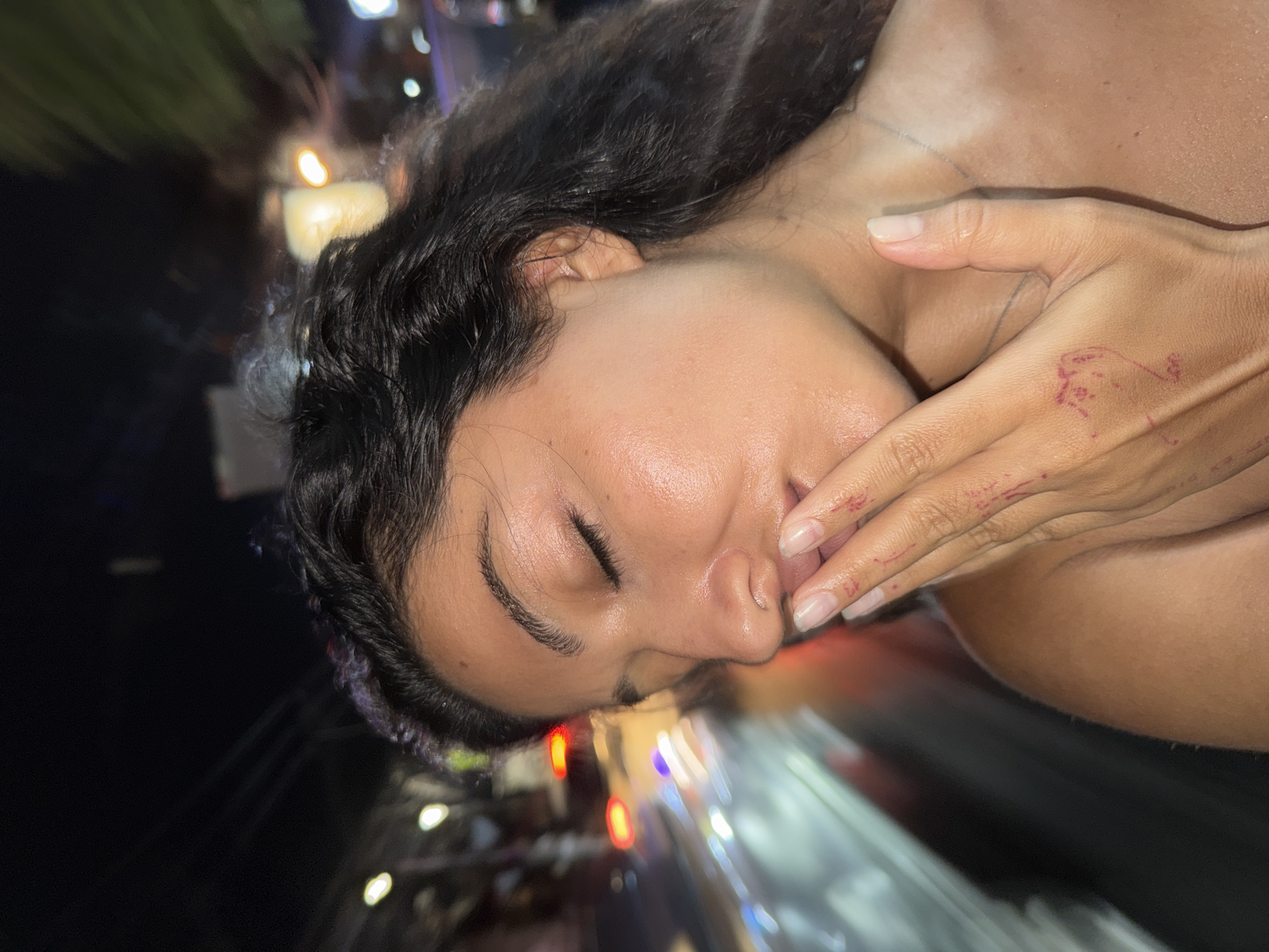 This close-up image shows a person, likely female, with dark, wavy hair and tan skin, positioned on their side with their head angled to the left. Their eyes are closed, and their right hand covers their mouth and lower nose. Red markings, possibly from temporary ink or paint, are visible on the back of the hand and fingers, which feature neatly trimmed, natural nails. A faint line, possibly a necklace, can be seen on the subject's neck and collarbone area.
The background is heavily out of focus, dominated by blurred streaks and bokeh effects from artificial lights in white, red, and orange hues against a dark setting. This suggests an outdoor, urban, or street environment at night. The motion blur in the background could indicate that the subject was in a moving vehicle or that the camera itself was moving during the capture. The overall impression is one of introspection, rest, or possibly exhaustion. There is no visible text in the image. The location context for the scene is North Kuta, Indonesia.