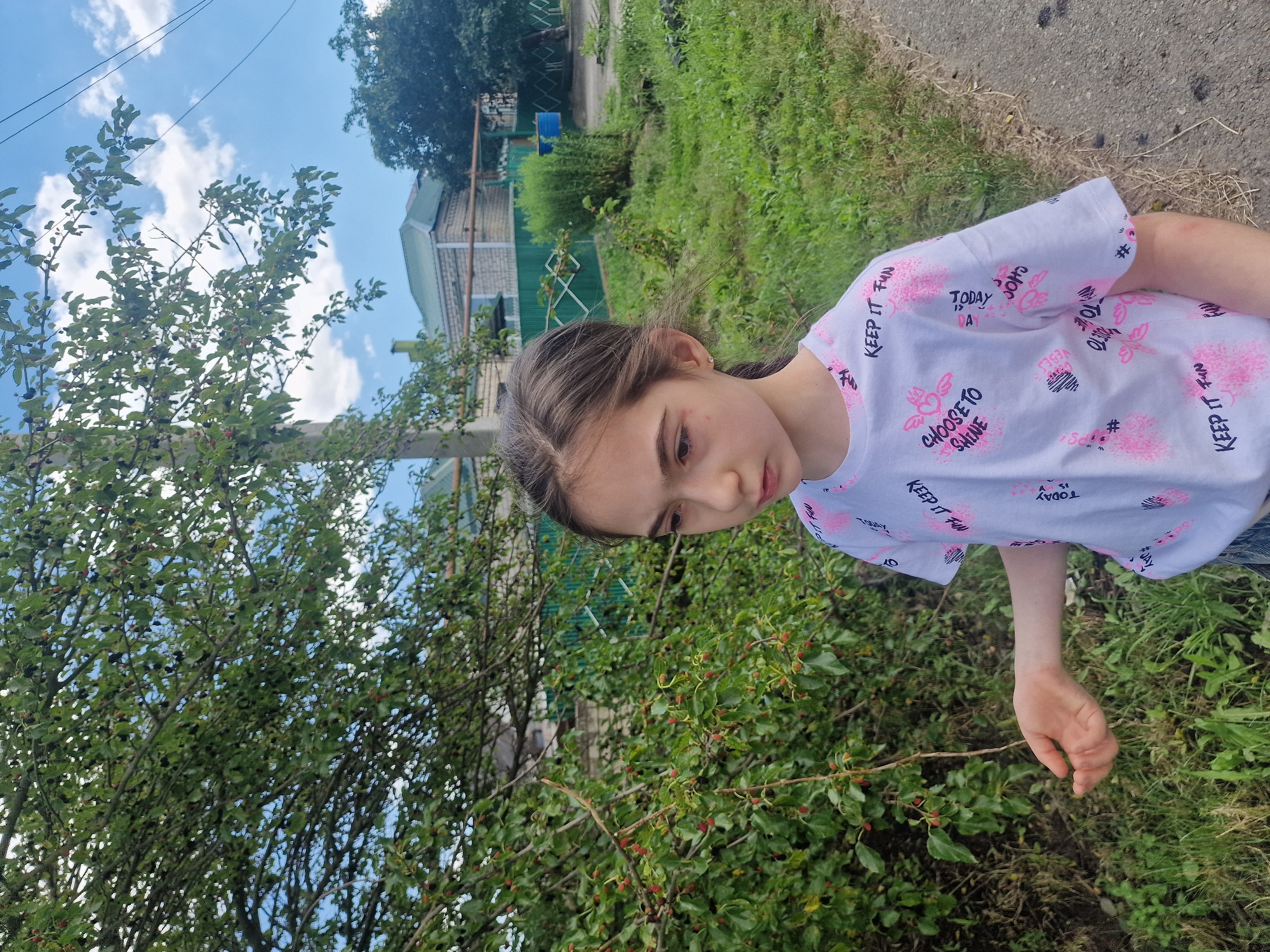 A young girl with dark hair pulled back stands outdoors, partially obscured by foliage from a mulberry tree. She is wearing a white t-shirt with pink text and graphics, including the phrases "KEEP IT FUN" and "CHOOSE TO SHINE." Her right hand is extended, palm open, as if reaching for something. The background depicts a rural or suburban setting in Kursavka, Russia, with a fence, buildings, and a dirt road. The sky is partly cloudy with a bright blue hue. The overall impression is of a casual, everyday moment in a residential area.
