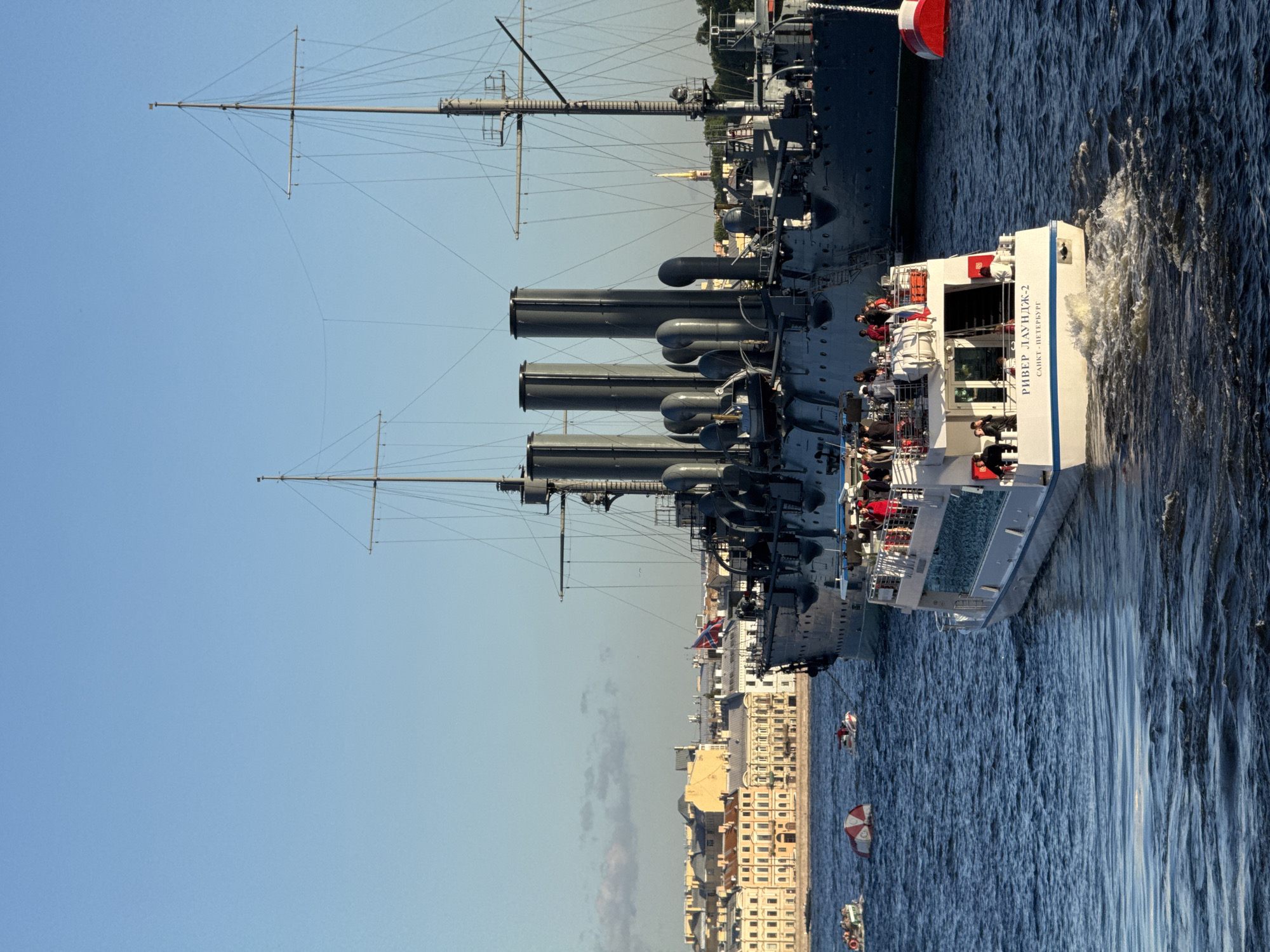 In the foreground of the image, a white passenger boat with a blue stripe travels on choppy blue water. The boat has a multi-level deck with people visible on both levels, some standing and some sitting. The text "РИВЕР ЛАУНДЖ-2" and "САНКТ-ПЕТЕРБУРГ" are visible in black lettering on the stern of the boat.
Behind the passenger boat, a large, dark gray ship, which appears to be a historical warship or museum ship, is docked. This ship has multiple smokestacks, masts, and rigging. The Russian flag is visible on its deck.
In the background, a cityscape with light-colored buildings lines the waterfront. The sky is clear and blue with a few faint clouds. Several smaller boats and buoys are also present in the water. The overall scene suggests a sunny day on a river or harbor.