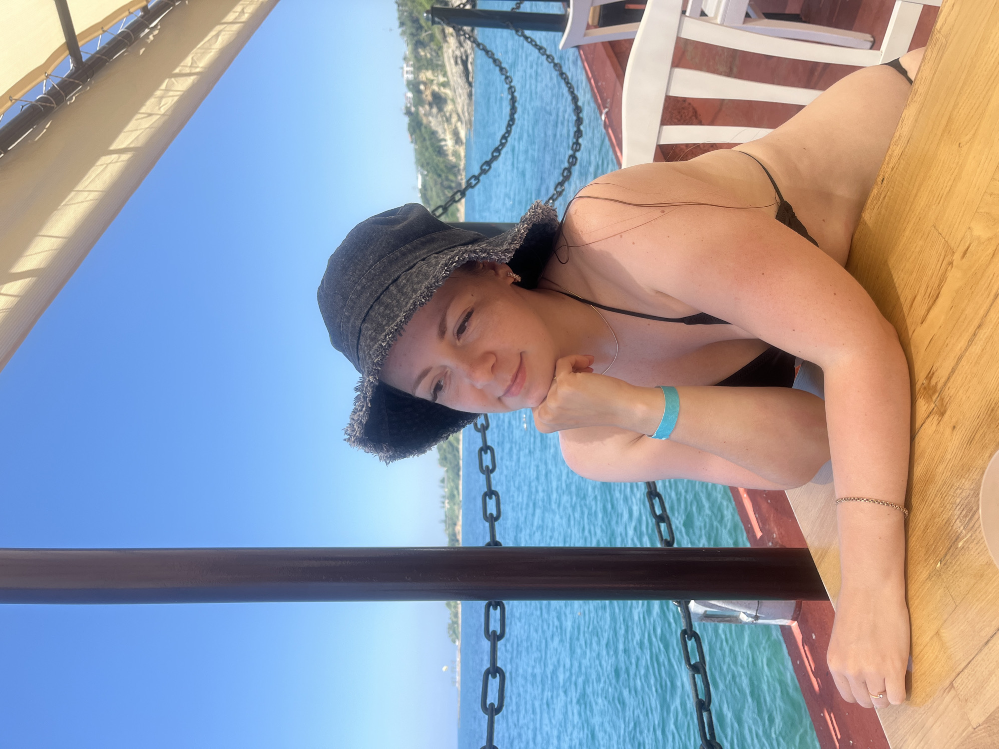 A woman wearing a black bucket hat and a black bikini is seated at a wooden table outdoors. She is looking to her left with her chin resting on her hand. A turquoise wristband is visible on her left wrist. In the background, the blue sea is visible, with a coastline and buildings in the distance. A dark metal chain runs along the edge of the seating area. The setting appears to be a cafe or restaurant overlooking the sea in Alanya, Turkey.