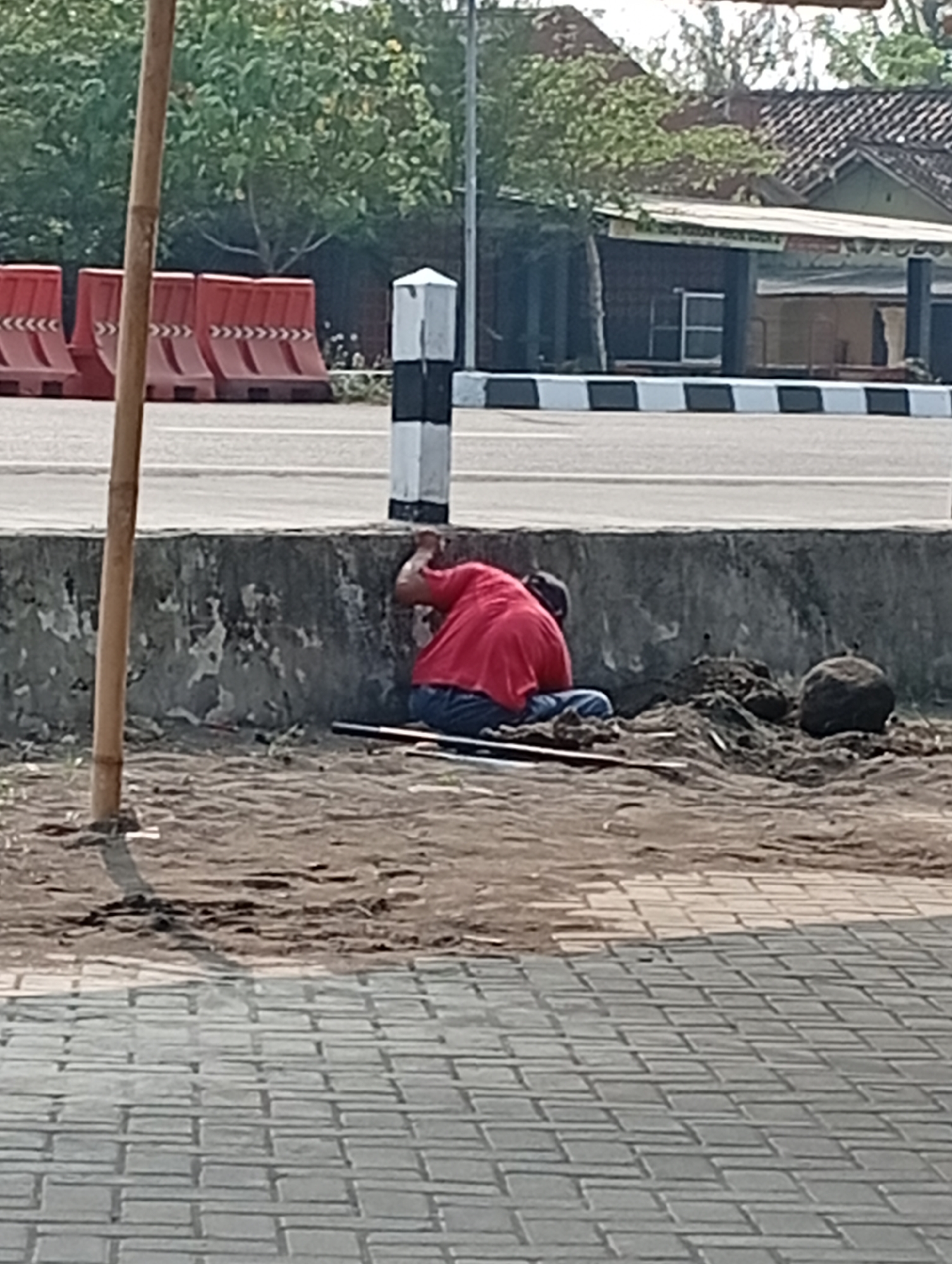 The image depicts an outdoor scene in Srandakan, Indonesia. In the foreground, a light-colored bamboo pole is visible on the left. Further back, a person wearing a red shirt and blue pants is crouching next to a concrete barrier, appearing to be engaged in some activity. Their head is down, obscuring their face. The ground around the person is a mixture of dirt and some debris, including what appears to be a black rod or pole. Behind the concrete barrier is a paved road with a black and white striped border. In the background, there are red traffic barriers, trees, and buildings with tiled roofs. The overall impression is an urban or semi-urban environment.