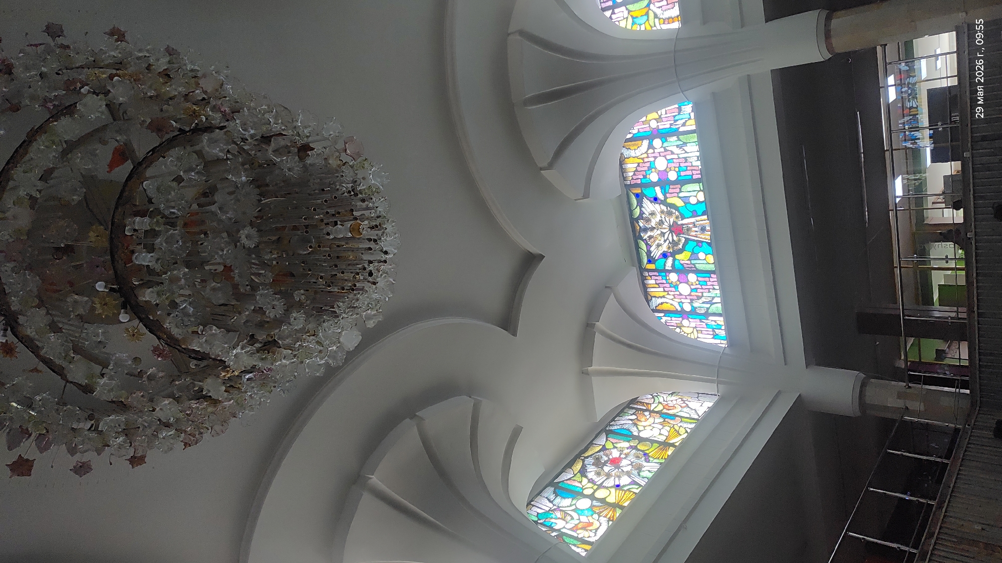 The image shows the interior of what appears to be a building in Astana, Kazakhstan, based on architectural style. The view is directed upward, focusing on an ornate chandelier, white vaulted ceilings with decorative molding, and stained-glass windows. The chandelier is complex, featuring multiple tiers and numerous small glass elements. The stained-glass windows exhibit colorful patterns and designs, with some depicting floral motifs. A structural column is visible in the upper-right portion of the image, along with a partial view of the building's interior, suggesting a mezzanine or balcony level. The date "29 May 2026, 09:55" is visible in the upper-right corner of the image. The environment is brightly lit due to natural light filtering through the stained-glass windows.
