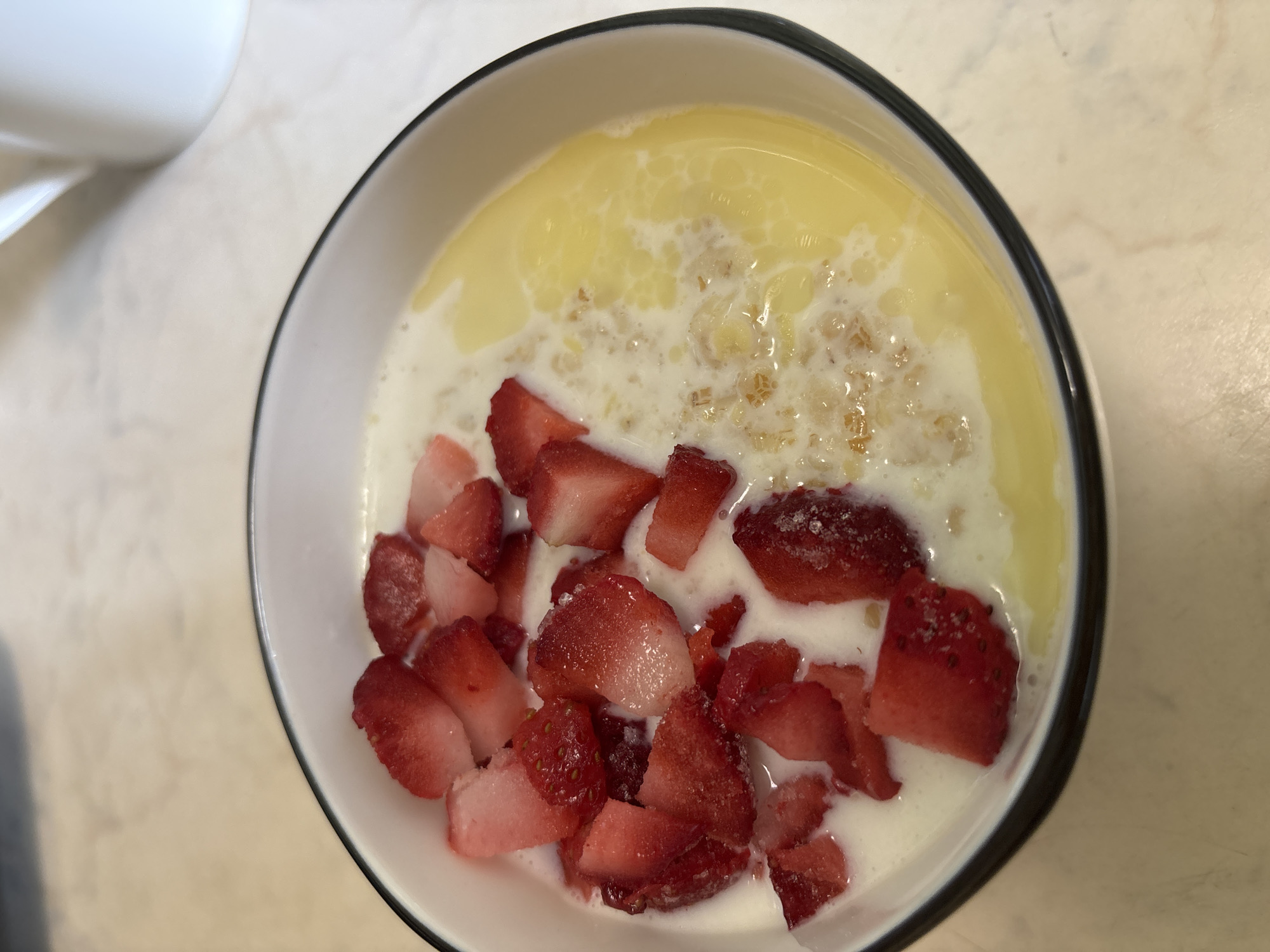 A close-up, overhead shot shows a white bowl with a black rim. Inside, diced red strawberries are piled on the left side. The right side contains a mix of white milk, yellow liquid, and what appears to be cereal flakes. A white mug is partially visible in the upper-left corner of the image. The bowl rests on a light-colored surface. The location is not discernable from the image.