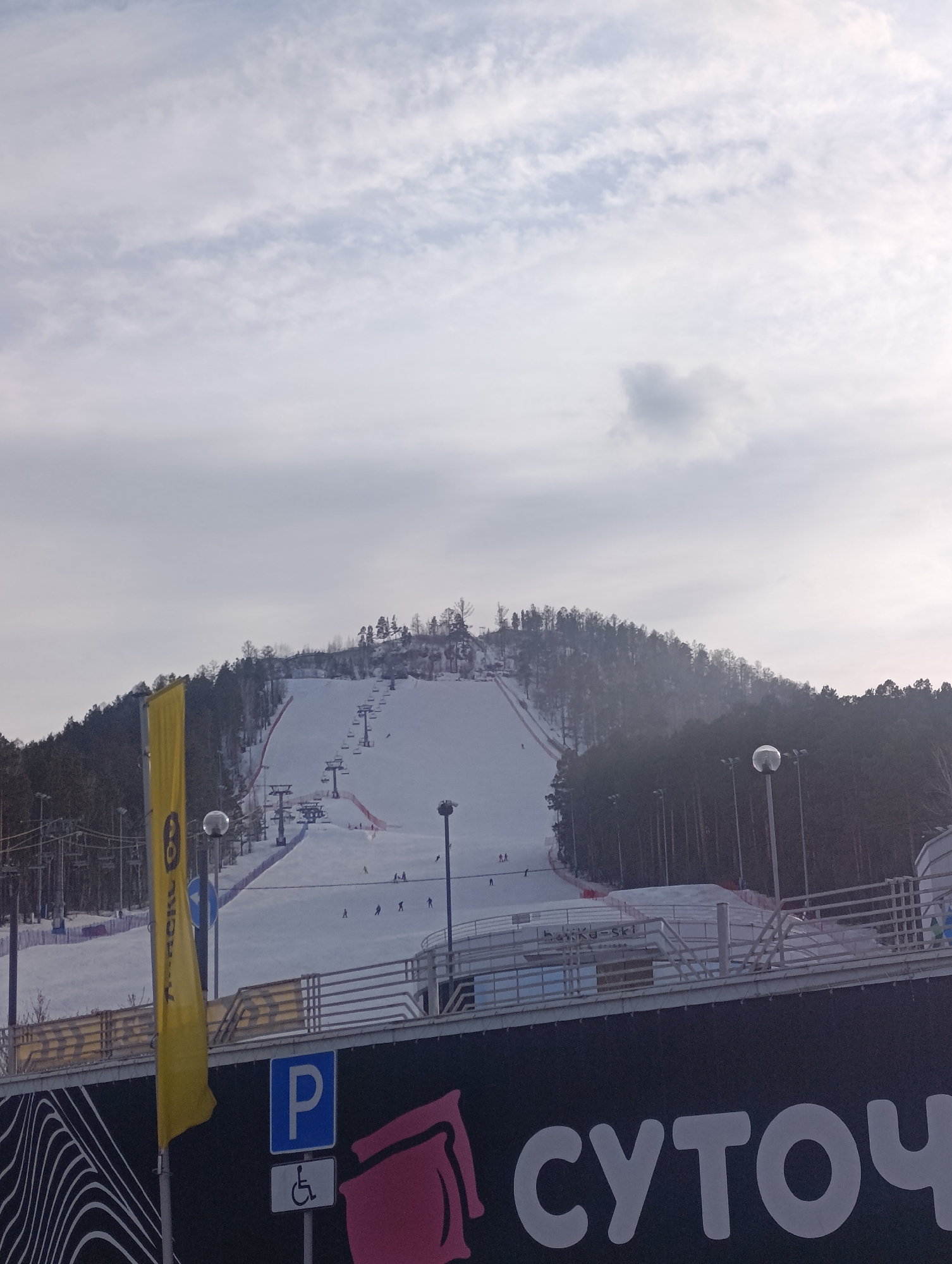 The image displays the Bobrovy Log Funpark ski resort in Krasnoyarsk, Russia. A snow-covered mountain, densely forested with coniferous trees, dominates the background under an overcast, light gray sky. Multiple ski slopes are visible, serviced by several multi-chair ski lift systems ascending the mountain. Approximately ten to fifteen individuals, appearing as skiers or snowboarders, are sparsely distributed across the slopes, some in descent. Red safety netting borders the left side of the main visible ski run. In the foreground, a prominent dark banner or wall features large white Cyrillic text, partially legible as "СУТОЧ" (SUTOCH). Below this text, a blue square sign with a white "P" indicates parking, accompanied by an international disabled access symbol. To the left, a tall yellow banner with dark text is attached to a pole, partially obscuring a blue circular road sign. A multi-story building structure with white railings is situated above the foreground banner, overlooking the ski slopes. Several streetlights with spherical luminaires are also visible within the scene.