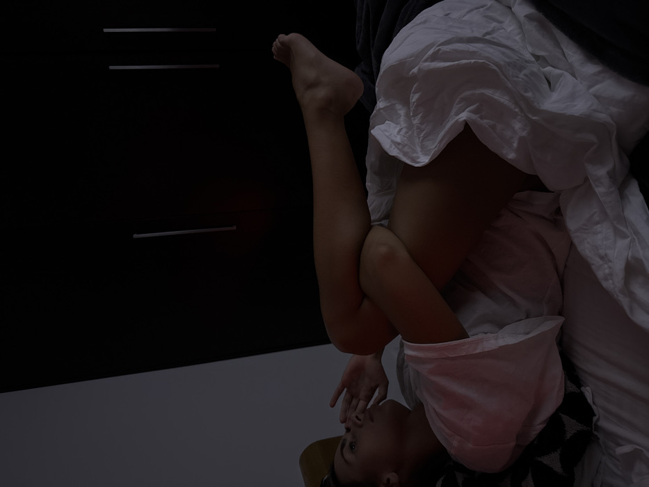 A dimly lit image features a person lying upside down on a patterned surface. Their legs are raised and bent, with a white garment draped over them. The person's face is visible, hand covering the mouth. In the background, there is a dark cabinet with thin, horizontal handles and a pale floor. The overall composition is moody, using the darkness to highlight the human form and the texture of the fabrics.