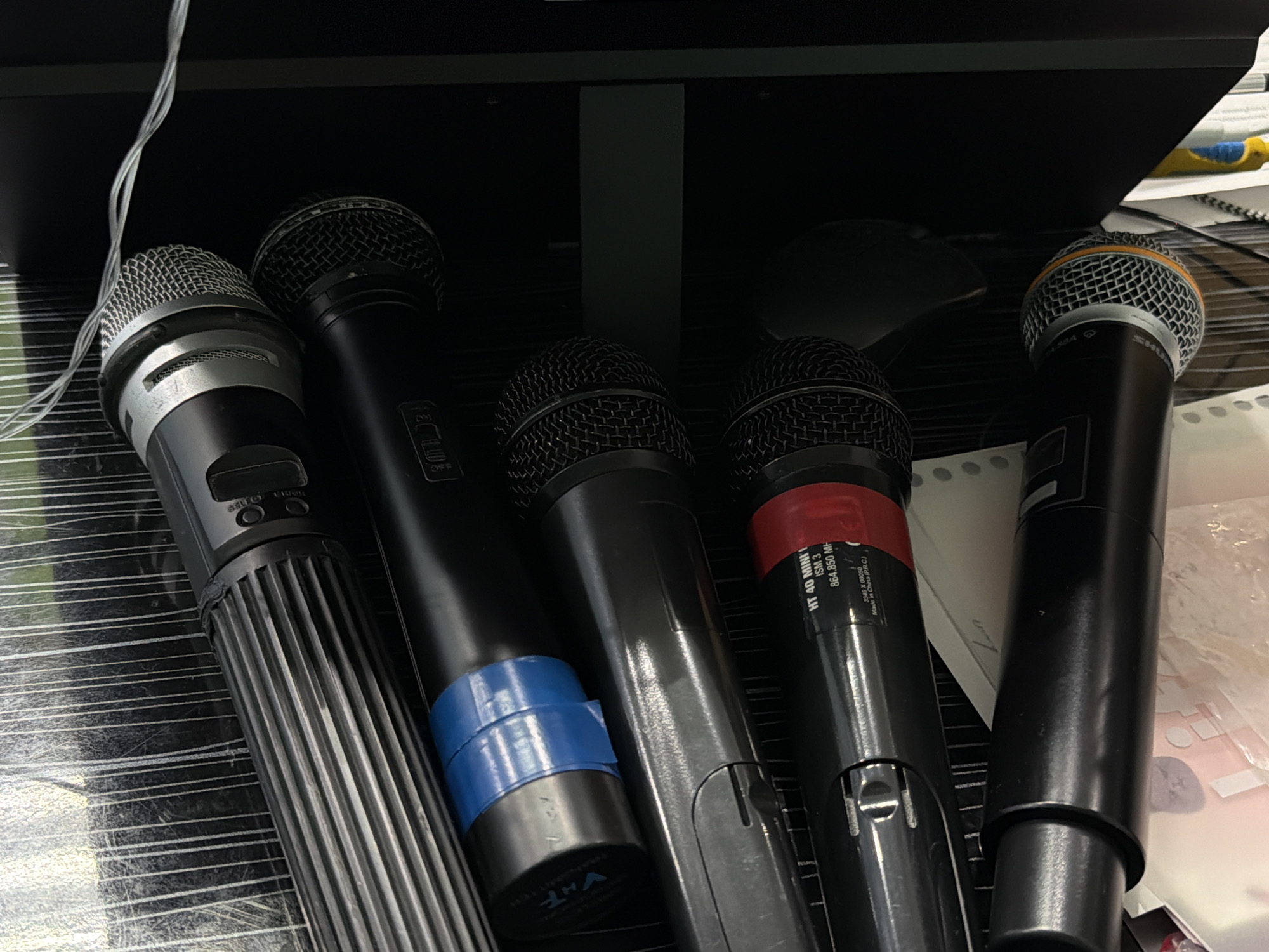 The image shows a close-up view of five handheld microphones placed on a surface, likely a desk or table, possibly located in Perm, Russia. The microphones vary in design, with one featuring a silver and black body with vertical ridges and visible control buttons. Another has a blue band wrapped around its lower portion. A third has a red band and labeling that includes the text "HT 40 MINII ISM 3 864.850 MH" and "Made in China (PRC)." All five microphones appear to be in a static, non-operational state, simply resting on the surface. The background is cluttered and suggests a workspace, with a computer monitor visible in the upper portion of the frame. Cables are seen in the upper left corner.