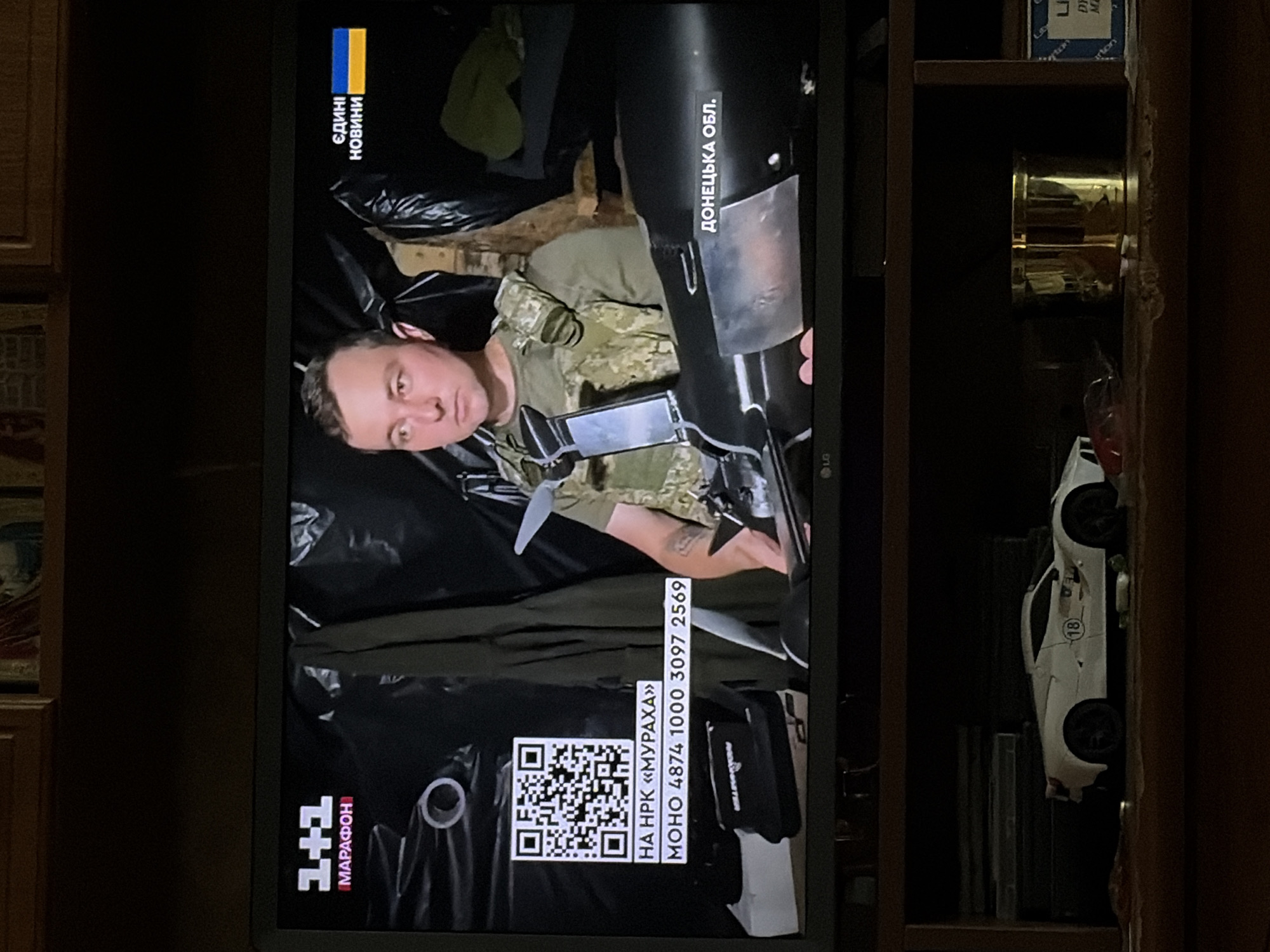 An image shows a television screen displaying a news broadcast. On the left side of the screen, the logo for "1+1 MARATHON" is visible, along with a QR code and text in Ukrainian. In the center of the screen, a man wearing a military-style vest is seated and holding a dark-colored object, possibly a weapon or equipment. To the right of the man, the Ukrainian flag and the words "ЄДИНІ НОВИНИ" (United News) are displayed. Below the man and the object, text in Ukrainian reads "ДОНЕЦЬКА ОБЛ." (Donetsk Oblast). The background of the broadcast appears to be dark and possibly industrial or makeshift. Below the television, a shelf holds a white toy car with the number "18" on its side, and a gold cylindrical object.