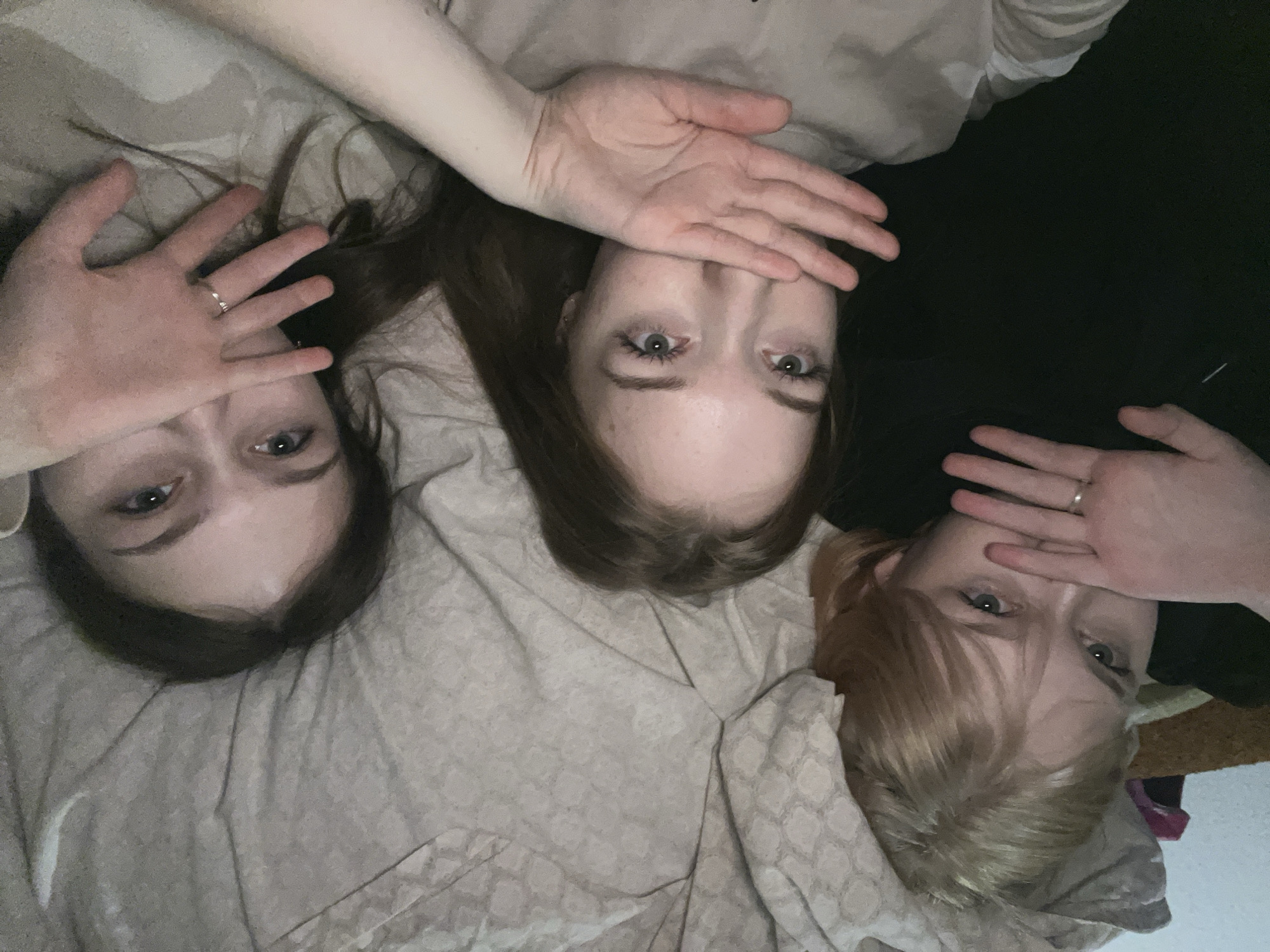 Three young women are lying side-by-side on a beige bedspread in a location that appears to be indoors. Each woman partially covers her face with her hand. From left to right, the first woman has dark brown hair, the second has medium brown hair, and the third has blonde hair. The second woman is positioned slightly higher in the frame compared to the other two. A dark, possibly black, fabric is seen between the second and third women. The location is possibly Chaikovsky, Russia, but there are no objects or environment to identify the location.