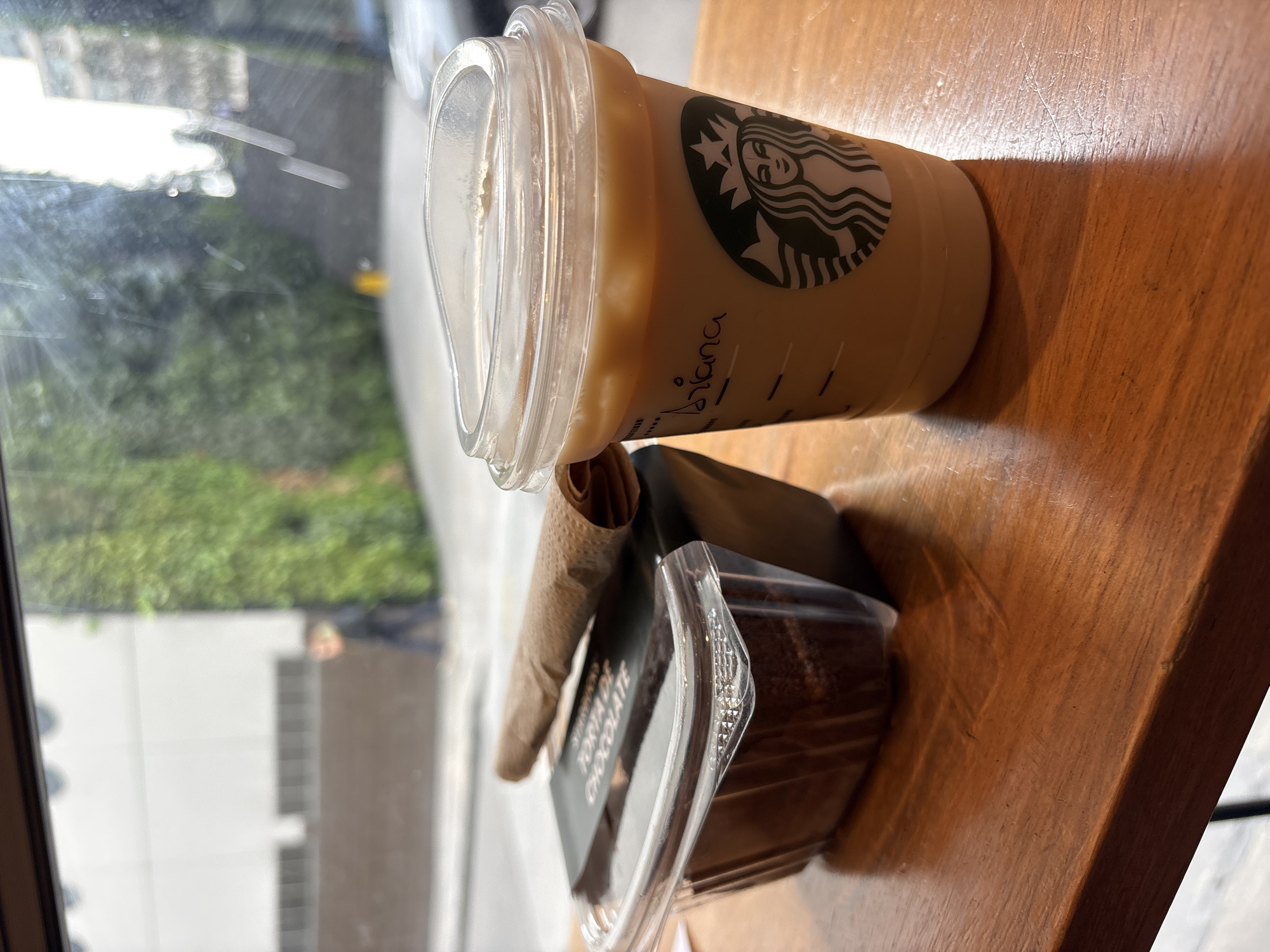 An iced coffee and a chocolate cake are on a wooden table. The coffee is in a clear plastic cup with a Starbucks logo and a handwritten name "Ariana" on it. The cake is in a clear plastic container with a black label that says "TORTA DE CHOCOLATE." A brown napkin is placed on top of the cake. In the background, through a window, a person is walking on a street. The area outside appears to be urban with buildings and greenery. The scene suggests a casual break or meal in a café or public space. The location is identified as San Isidro, Peru.