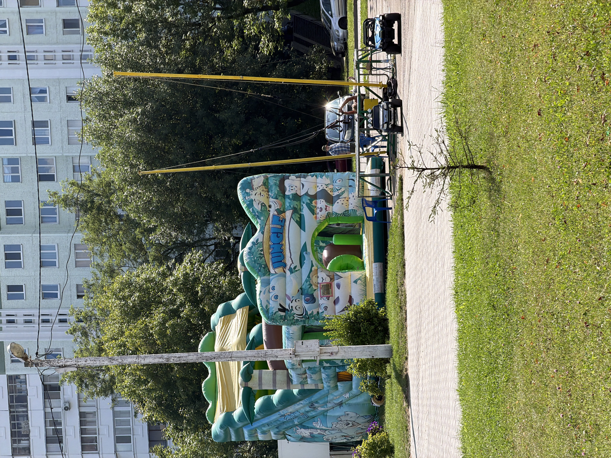 In a paved area adjacent to a grassy lawn, a large, inflatable jungle-themed bounce house with a slide and archway is set up. A lamppost stands to the left of the bounce house. Behind the bounce house, trees and a multi-story apartment building with light-colored facades and numerous windows are visible. To the right of the bounce house, a bungee trampoline attraction is in operation with two yellow poles and a platform, where a person in a blue and white checkered shirt appears to be participating. Two small, electric toy vehicles, one black and one blue, are parked near the bungee trampoline platform. Several cars are parked further in the background, including a silver sedan and a darker colored SUV. The scene is illuminated by bright daylight.