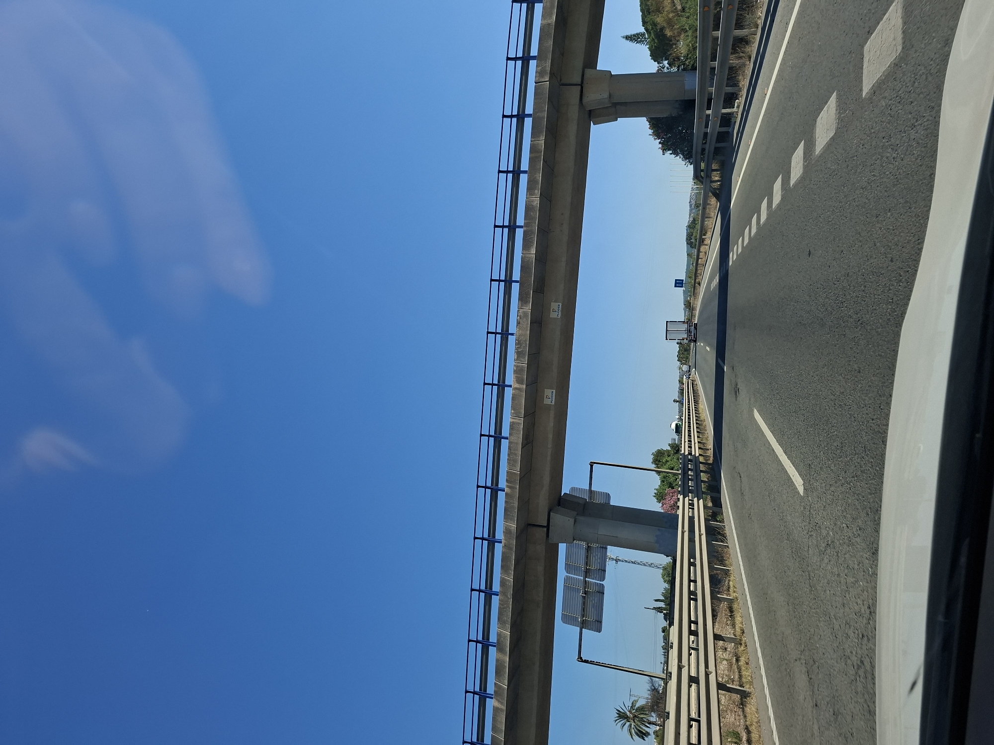 This is a daytime, eye-level, exterior shot taken from a moving vehicle on a highway. The highway has multiple lanes marked with white lines. In the foreground, the hood of a white vehicle is visible. An elevated highway overpass with blue railings runs across the frame, supported by concrete pillars. There are signs on the overpass and a billboard on a concrete pillar on the left. Palm trees and other vegetation are present on the sides of the highway, along with guardrails. A truck and other vehicles are visible on the highway in the distance, under a clear blue sky with some wispy clouds. The scene is set in Almoradí, Spain.